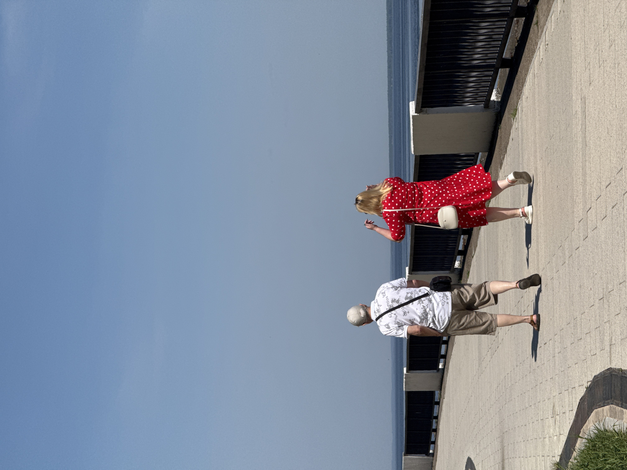On a sunny day in an unknown city, two people, a woman and a man, stand on a paved walkway bordering the sea or a large lake. The walkway is separated from the water by a dark metal railing. The woman, positioned further along the walkway, wears a red dress with white polka dots and white sneakers and carries a small white purse. Her blonde hair is down. She has one arm raised. The man, walking behind her, is dressed in a light-colored, patterned short-sleeved shirt, khaki shorts, and sandals. He wears a light-colored baseball cap and has a black bag slung across his body. The sky is a clear, light blue.