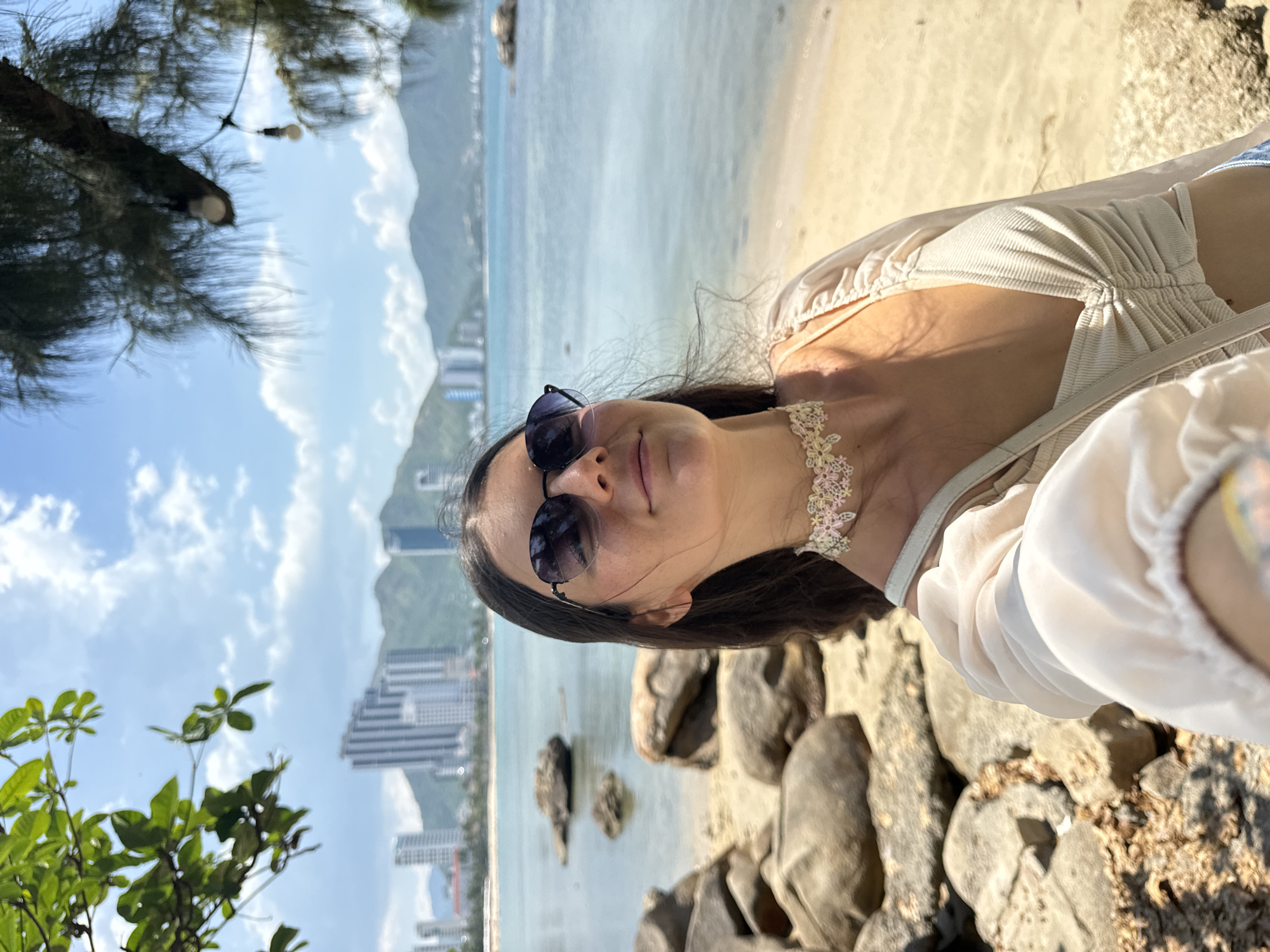 A woman takes a selfie on a beach in Nha Trang, Vietnam, with the ocean, distant buildings, and mountains visible in the background. She is wearing sunglasses, a light-colored top with ruching, and a floral choker necklace. Rocks and sand are in the foreground, and a tree with hanging lights is to the right, with leaves framing the top left corner. The sky is blue with scattered clouds.