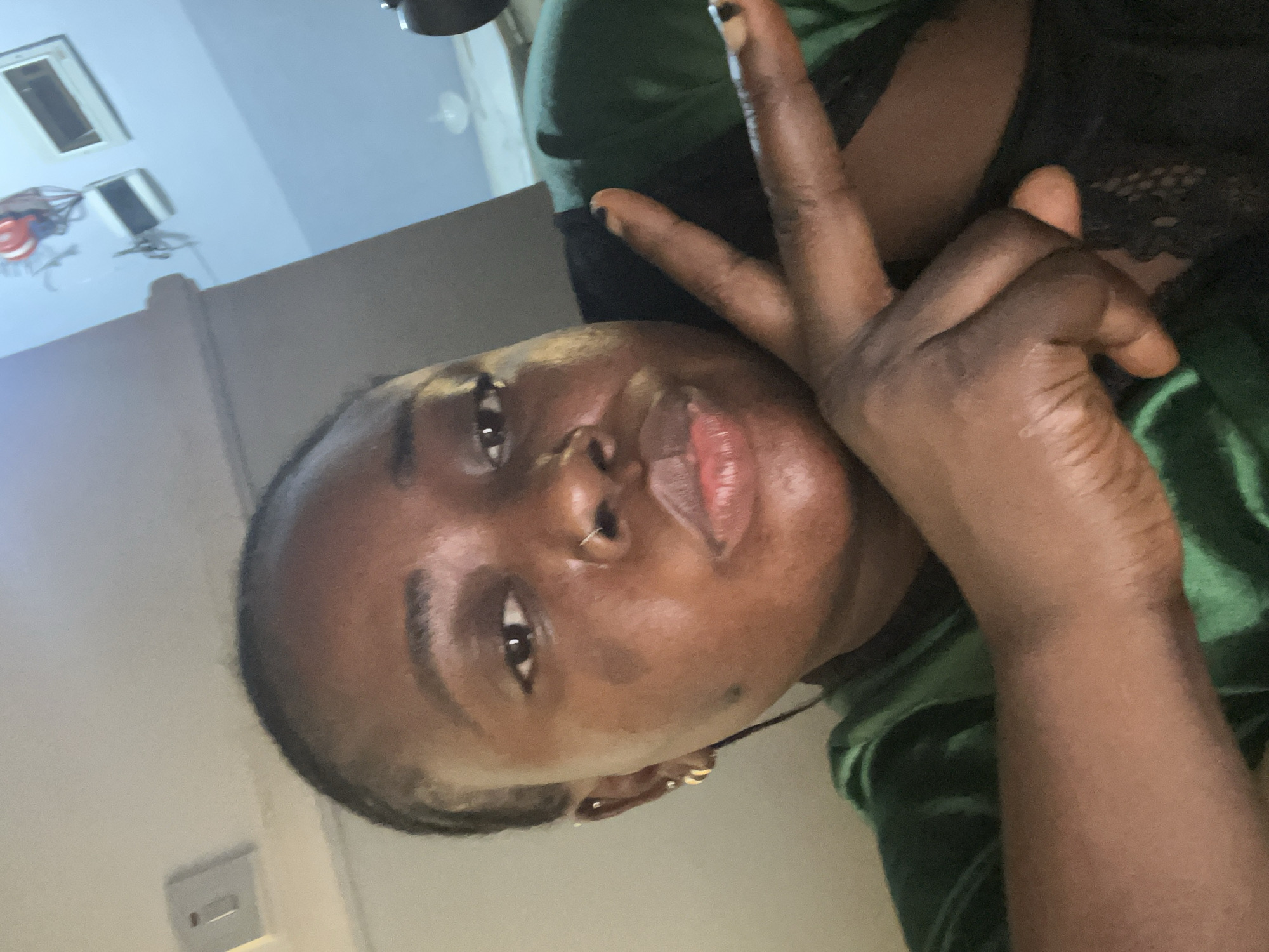 A dark-skinned person with short, dark hair is posing for a selfie. They are making a peace sign with their left hand, which is positioned near their face. They are wearing a green velvet-like garment. The background shows a room with a light-colored wall and what appears to be a light switch. A portion of the ceiling and a hanging object are also visible. The location of the image cannot be determined to be Kubwa, Nigeria based on the contents of the image provided.