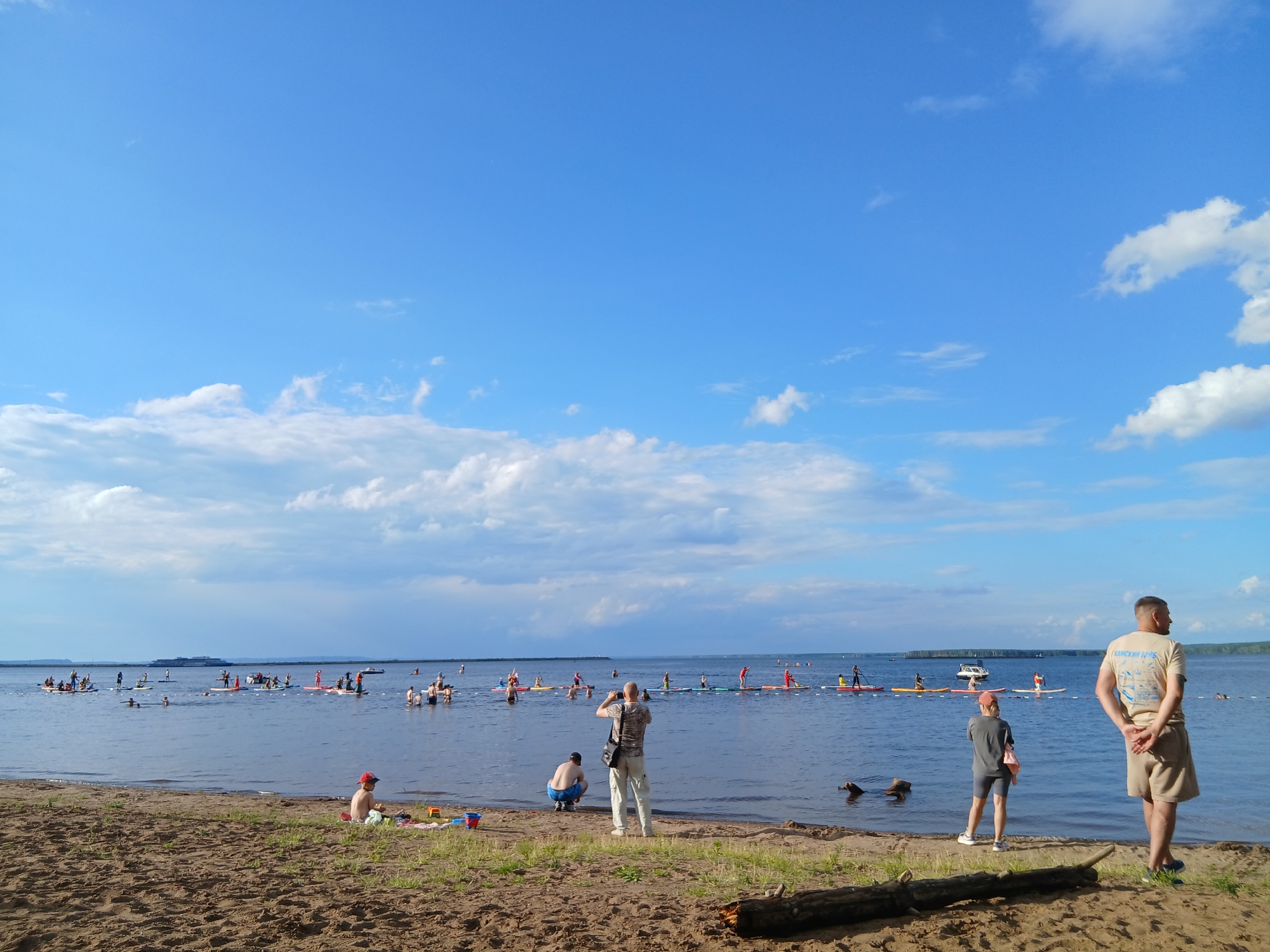 The image depicts a sunny day at a beach in Chaikovsky, Russia, with a clear blue sky and scattered white clouds. The scene is set by the water, likely a river or lake, where numerous people are engaged in stand-up paddleboarding. A long line of paddleboarders stretches across the water, with some individuals appearing to be part of an organized event. In the foreground, the sandy beach is visible, with a few people enjoying the shore.
On the beach, a young child wearing a red cap sits near the water's edge, playing with sand toys. Further back, two men are standing on the sand, one of whom is taking a photo with a camera. To the right, another man stands with his back to the camera, looking out at the water. The presence of multiple people on paddleboards suggests a recreational or sporting event. The overall atmosphere appears relaxed and enjoyable, indicative of a summer day.
Notable details include the bright sunshine, implying it is likely daytime, and the clear weather. The distant shoreline and a large ship or ferry in the background provide a sense of the location's scale. There is some visible text on the t-shirt of the man on the right, but it is not clearly legible in the image.