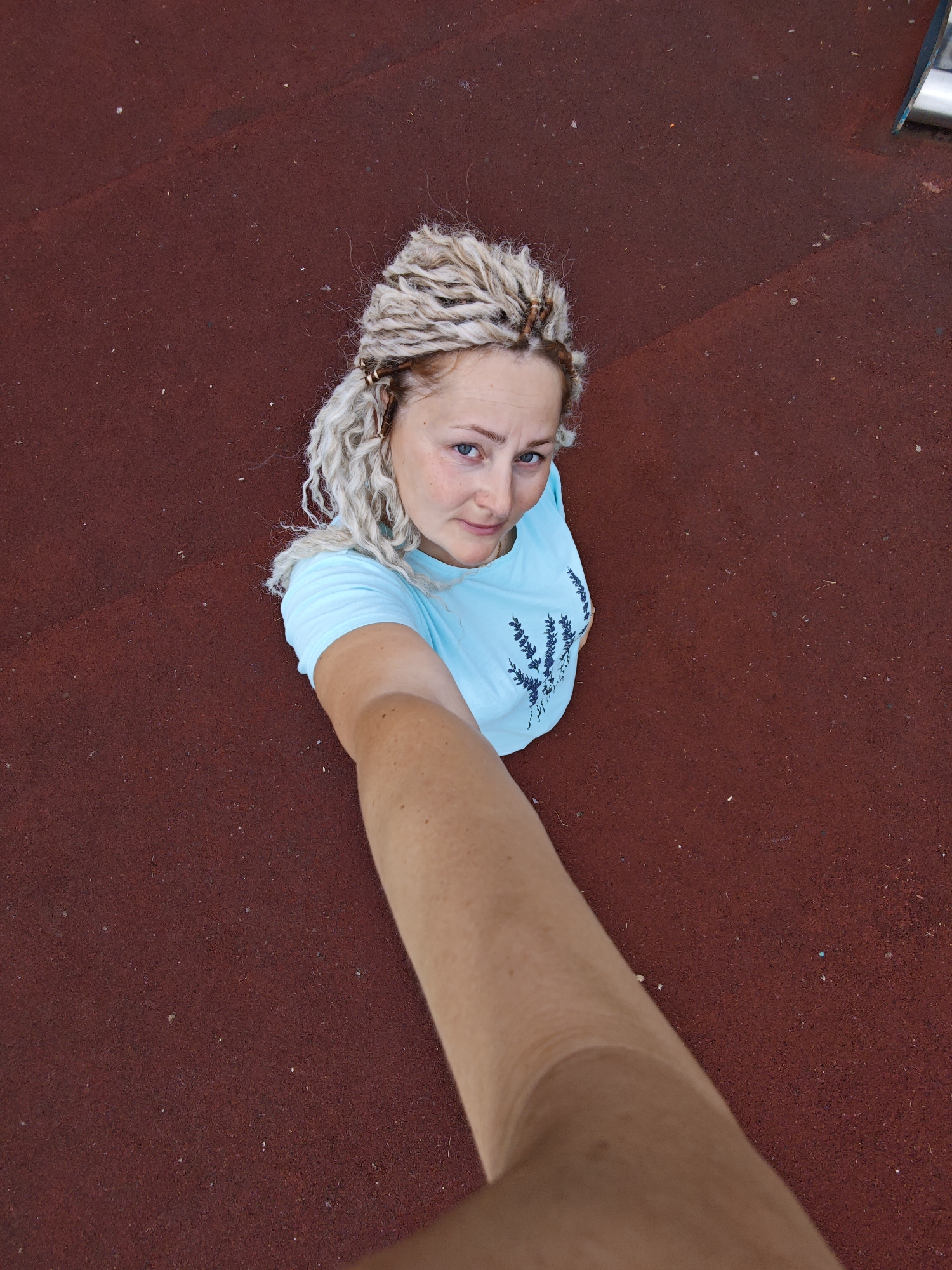 A woman is taking a selfie outdoors, standing on a dark red, textured surface. She has blonde hair styled in many small braids with some strands pulled back from her face. She is wearing a light blue t-shirt with a dark blue floral or plant-like design on the front. Her expression is neutral, and she is looking towards the camera. The background is the same red surface, which appears to be a sports or playground area, possibly with some subtle variations in color and texture. In the upper right corner, a small section of a metallic structure, possibly part of playground equipment, is visible. The lighting suggests it is daytime, and the weather appears to be clear. The overall scene is casual and appears to be a personal moment captured by the subject. There is no visible text in the image.