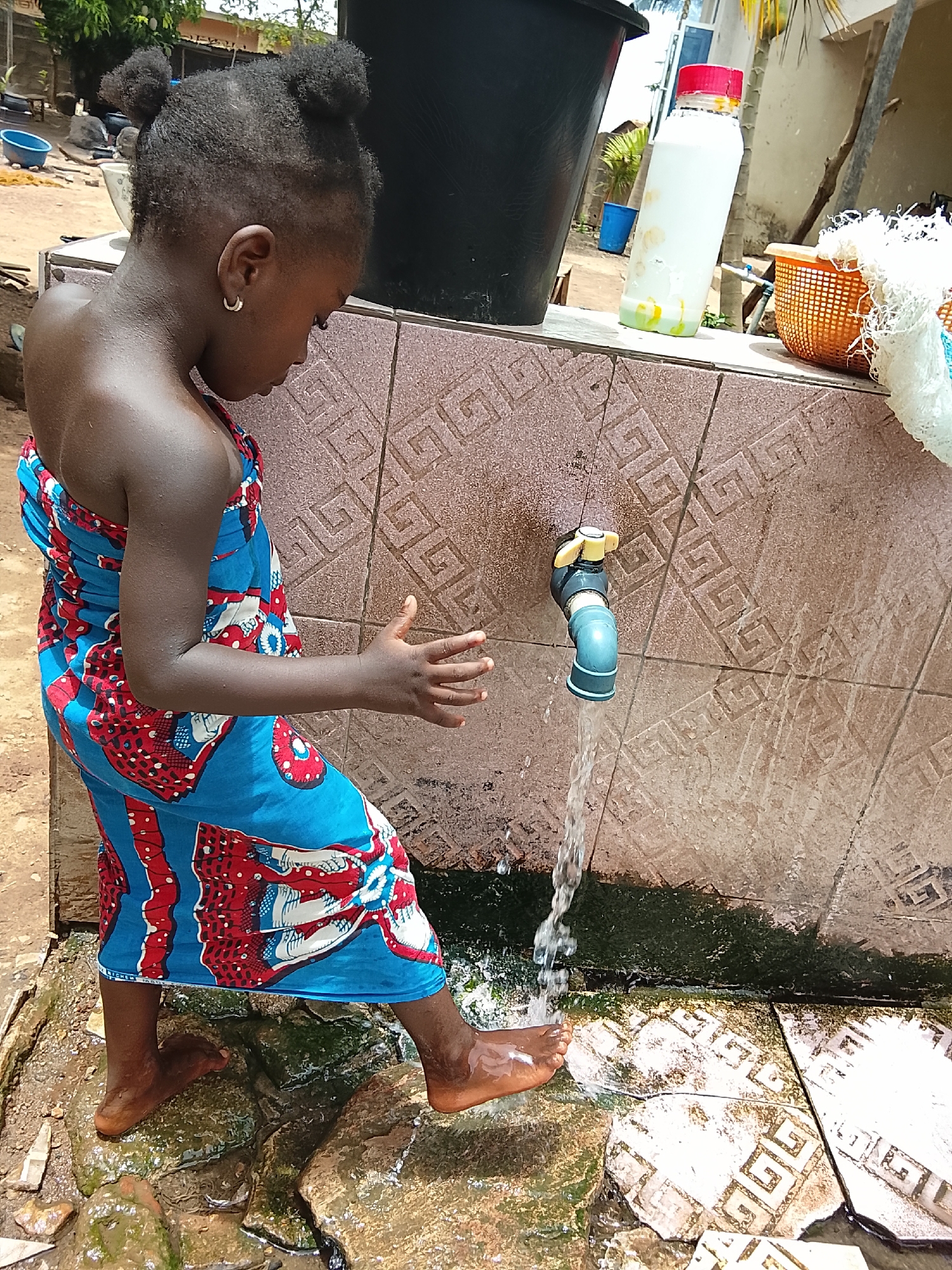 A young girl with braided hair styled in two buns, wearing a blue and red patterned wrap dress, stands at an outdoor tap in Akwanga, Nigeria. She is facing the tap and putting her hands and bare feet in the running water. The tap is attached to a tiled wall with a geometric pattern. Algae growth is visible around the base of the tap and on the tiled surface below. Behind the girl and the tap are a black bucket, a plastic bottle with a red lid, and an orange basket covered with white fabric. The ground is wet and appears to be tiled with a similar geometric pattern to the wall. The environment suggests a domestic setting, possibly a backyard or communal water access point.