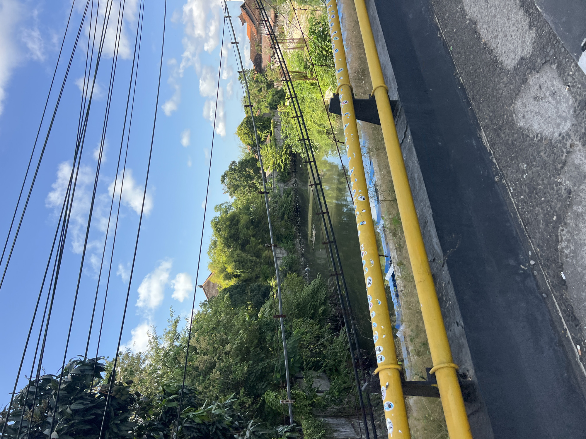 A view of a small river with overhanging trees and buildings in Kuta, Indonesia. The river is bordered by concrete walls and lush vegetation. In the foreground, a yellow railing with stickers on it is visible, suggesting it is part of a bridge or walkway. Several power lines crisscross the sky. The sky is blue with scattered white clouds. The overall impression is a semi-urban environment with a blend of natural elements.
