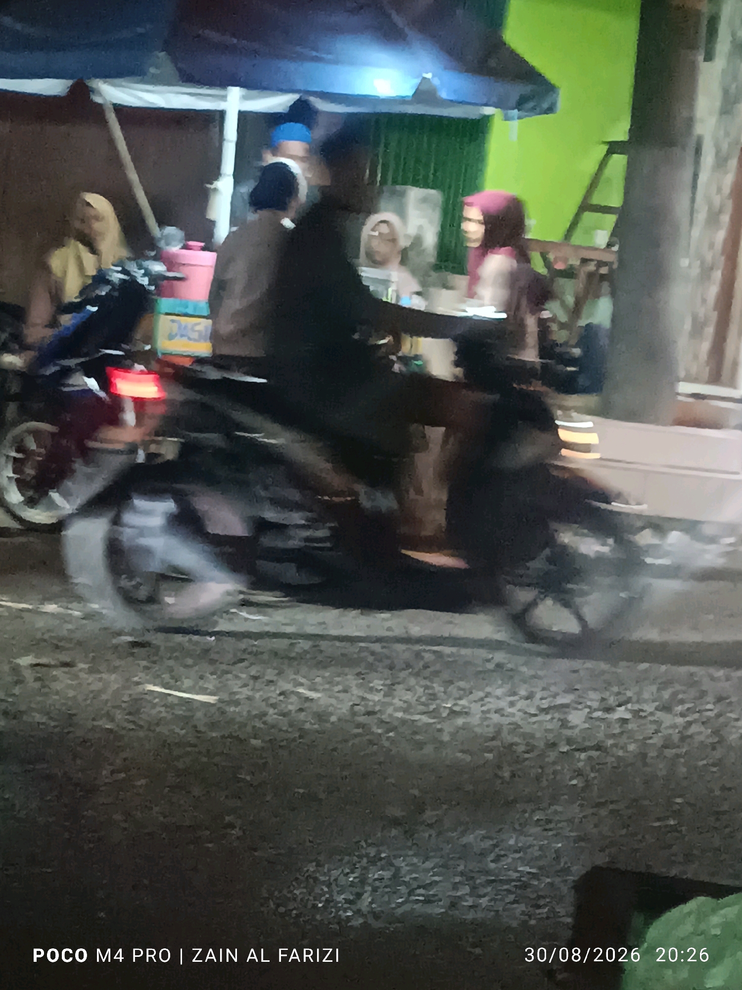 This is a blurry, low-light, outdoor photograph taken at night in the city of Buduran, Indonesia. A person is riding a dark-colored motorcycle with a red taillight visible. The motorcycle has a license plate that appears to read "DAS". Behind the motorcycle, there are several individuals and what looks like a food stall with a pink container. The background is indistinct, but suggests an urban environment with a building and greenery. The timestamp at the bottom of the image indicates it was taken on August 30, 2026, at 20:26.