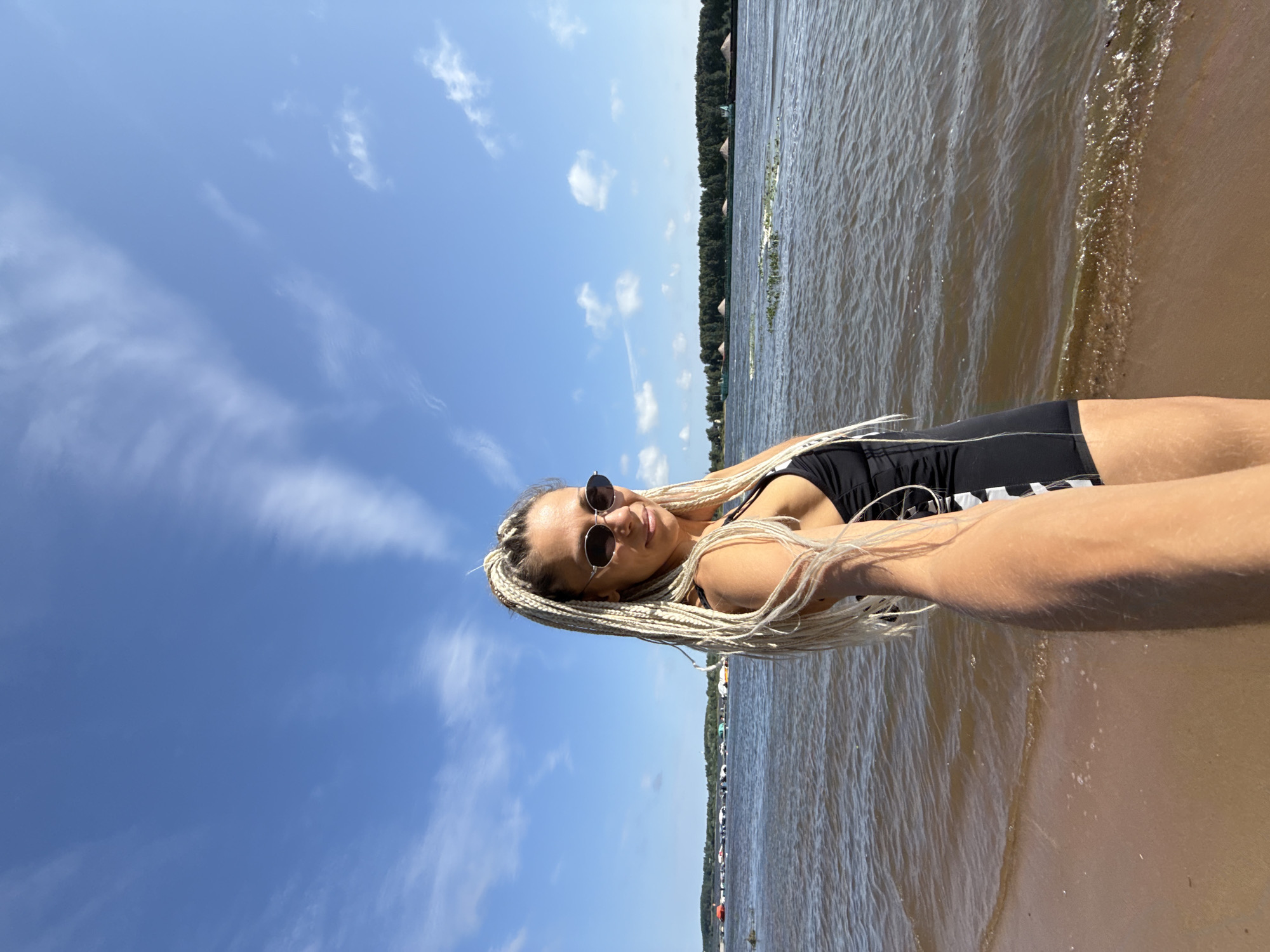 A young Caucasian woman with long, blonde braided hair, wearing a black swimsuit with white stripes and oval sunglasses, stands on a sandy beach next to a body of water. She is holding the camera, taking a selfie. In the background, there is a calm body of water with several small boats docked along the distant shore. A line of trees borders the far bank, and the sky above is a clear blue with scattered white clouds. The image appears to be taken on a sunny day. The location is the city of Dumino, Russia.