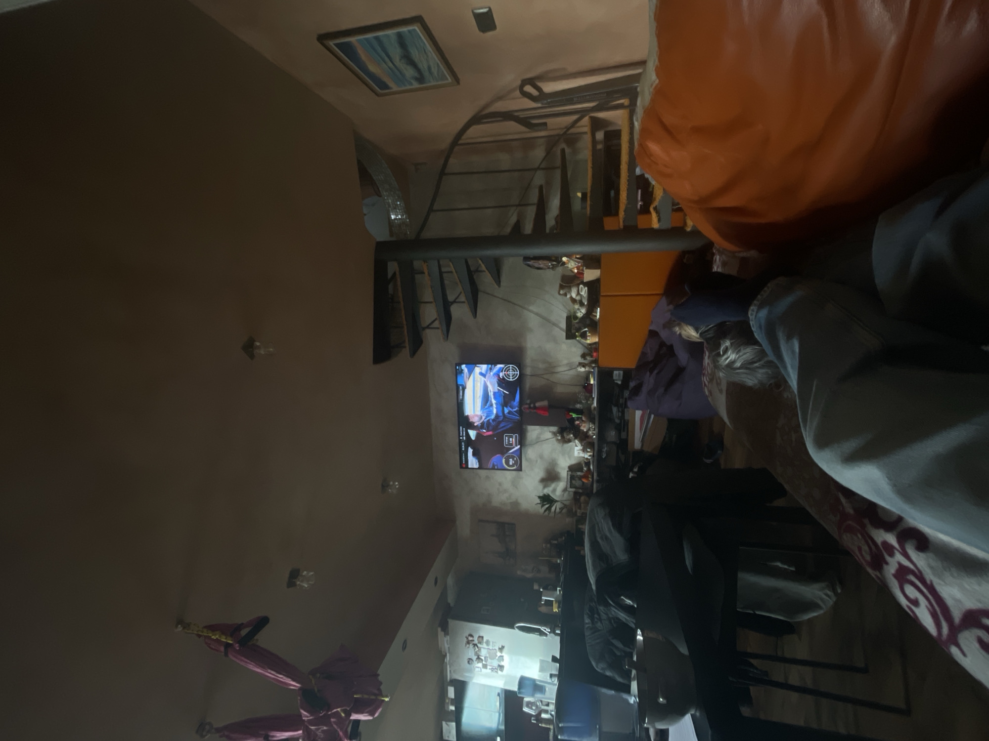 This image depicts an indoor scene, likely a living room or communal area, within a home in Burgas, Bulgaria. The perspective is from a low angle, showing a lived-in space with various personal touches.
In the foreground, on the right side, a person is visible, partially obscured by a large, orange, upholstered sofa or beanbag chair. Their legs, clad in jeans and dark socks, are prominent, suggesting they are lounging or relaxing.
The midground features a wall-mounted television screen, which is actively displaying what appears to be a video game, possibly a racing or action game, indicated by the dynamic graphics and a visible target reticle in the center. Below the television, shelving units hold an assortment of items, including footwear and decorative pieces. To the left of the TV, a prominent dark-colored spiral staircase with metal railings curves upwards, adding a unique architectural element to the room. Above the staircase, a framed artwork or picture hangs on the wall, and multiple light fixtures are visible on the ceiling and walls throughout the space.
In the background, further to the left, another area of the house, possibly a kitchen or dining space, can be seen, indicated by a refrigerator covered with magnets and other household items. On the far left of the frame, near the bottom, several folded pink or purple fabric objects, resembling umbrellas or decorative hangings, are suspended from the ceiling.
The overall lighting suggests evening or nighttime, relying on interior light sources. The walls are a warm, neutral shade. The scene gives the impression of a comfortable, casual environment where someone is engaged in leisure activity. No clearly legible text is discernible on any objects other than the general user interface elements on the television screen.