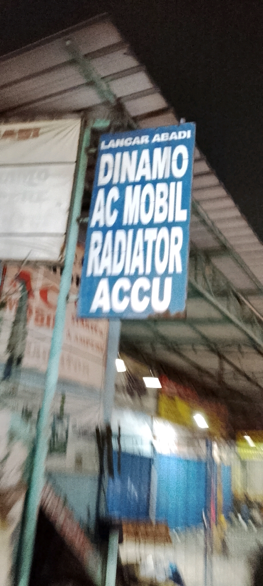 The image shows a blue sign with white text advertising car services. The text reads "LANGAR ABADI DINAMO AC MOBIL RADIATOR ACCU". The sign is hanging from a pole in what appears to be an outdoor market or workshop setting. In the background, there are buildings with corrugated metal roofs and some blurry figures. The overall scene suggests a commercial area. The provided location information indicates this is in Gunung Putri, Indonesia.