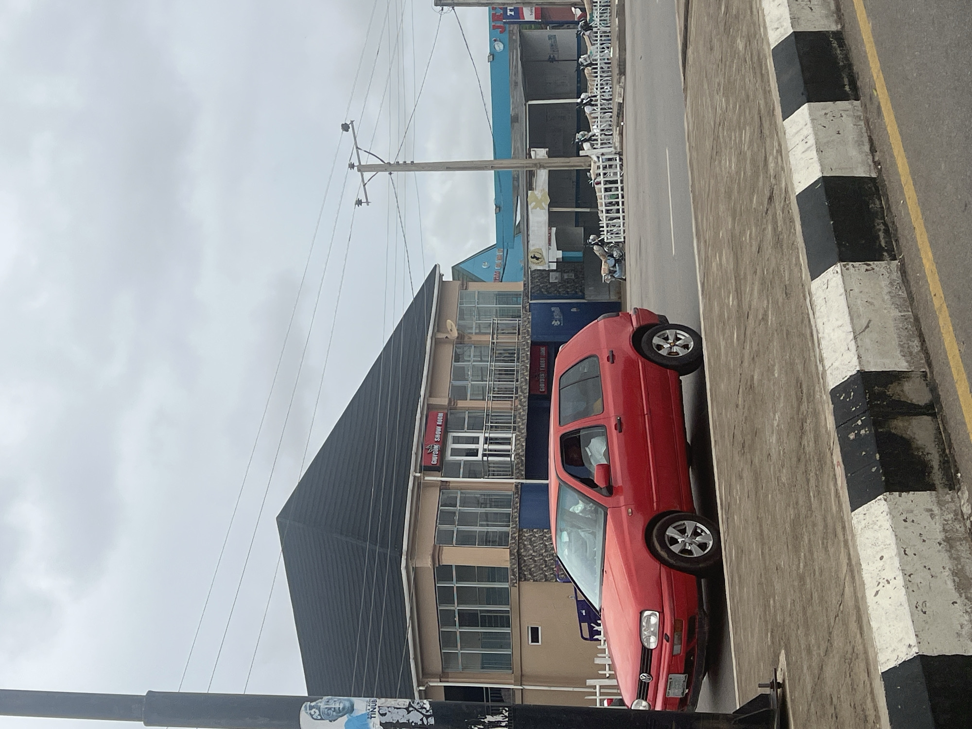 In Jalingo, Nigeria, a red Volkswagen Golf hatchback is parked on the side of a road, facing right. The car is a three-door model, and the driver's side window is down. The car is positioned on a paved area next to a sidewalk with black and white painted concrete barriers. Across the street, there is a building with a dark-tiled, peaked roof and large glass windows. A sign above the windows reads "GIOTOUK SHOW ROOM". Further down the street, a blue building is visible. Several motorcycles are parked in front of the blue building behind a white fence. A utility pole with wires extends across the sky. A black lamppost with posters affixed to it stands to the left of the car. The sky is overcast.