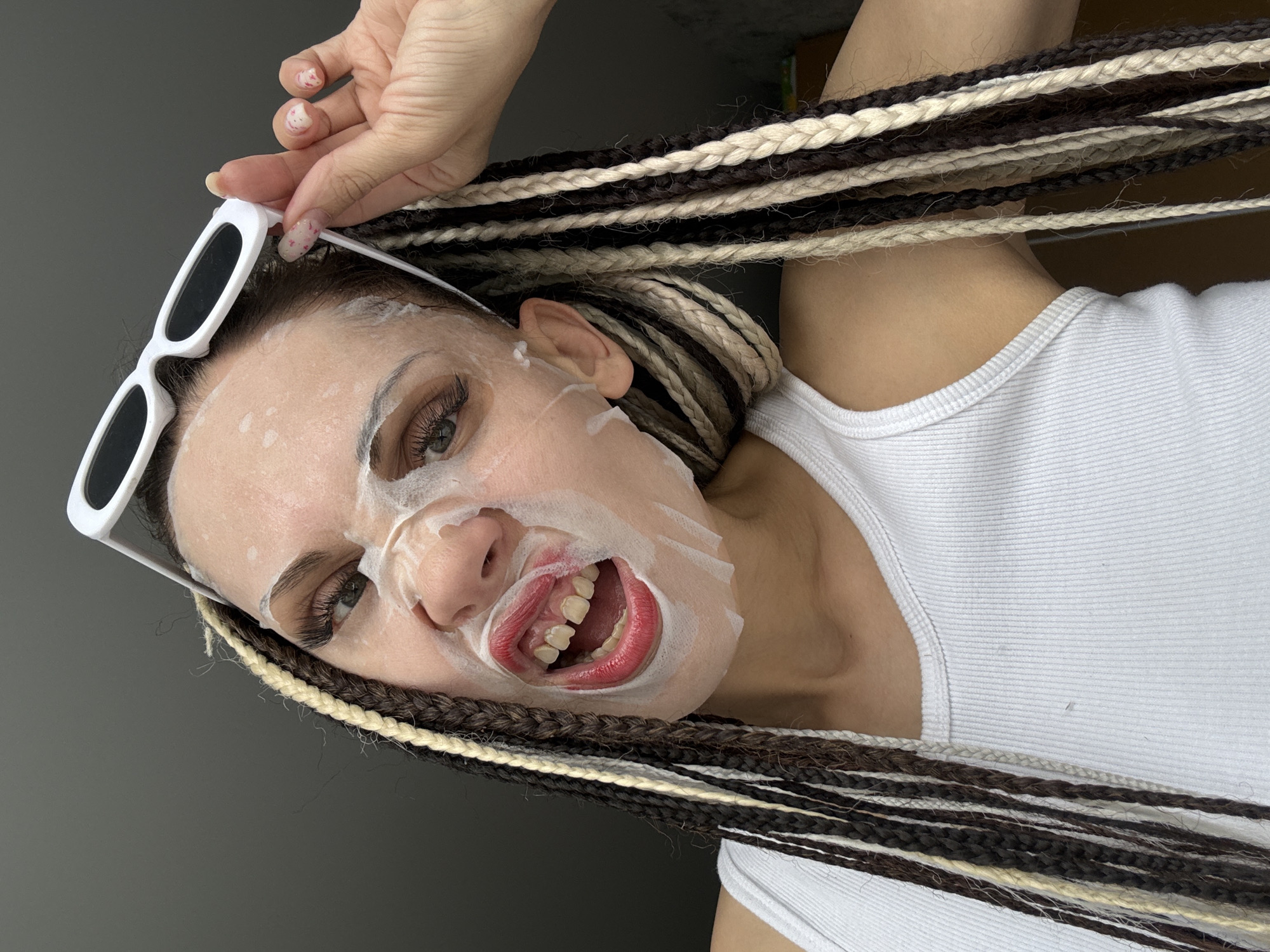 A woman with light skin is wearing a white face mask and a white tank top. She has long brown, beige, and white braided hair. She is holding white sunglasses on top of her head. She is looking at the camera with her mouth open, revealing her teeth. The background is a solid gray color. The image appears to be taken indoors and is angled diagonally. The location is not discernible from the image.