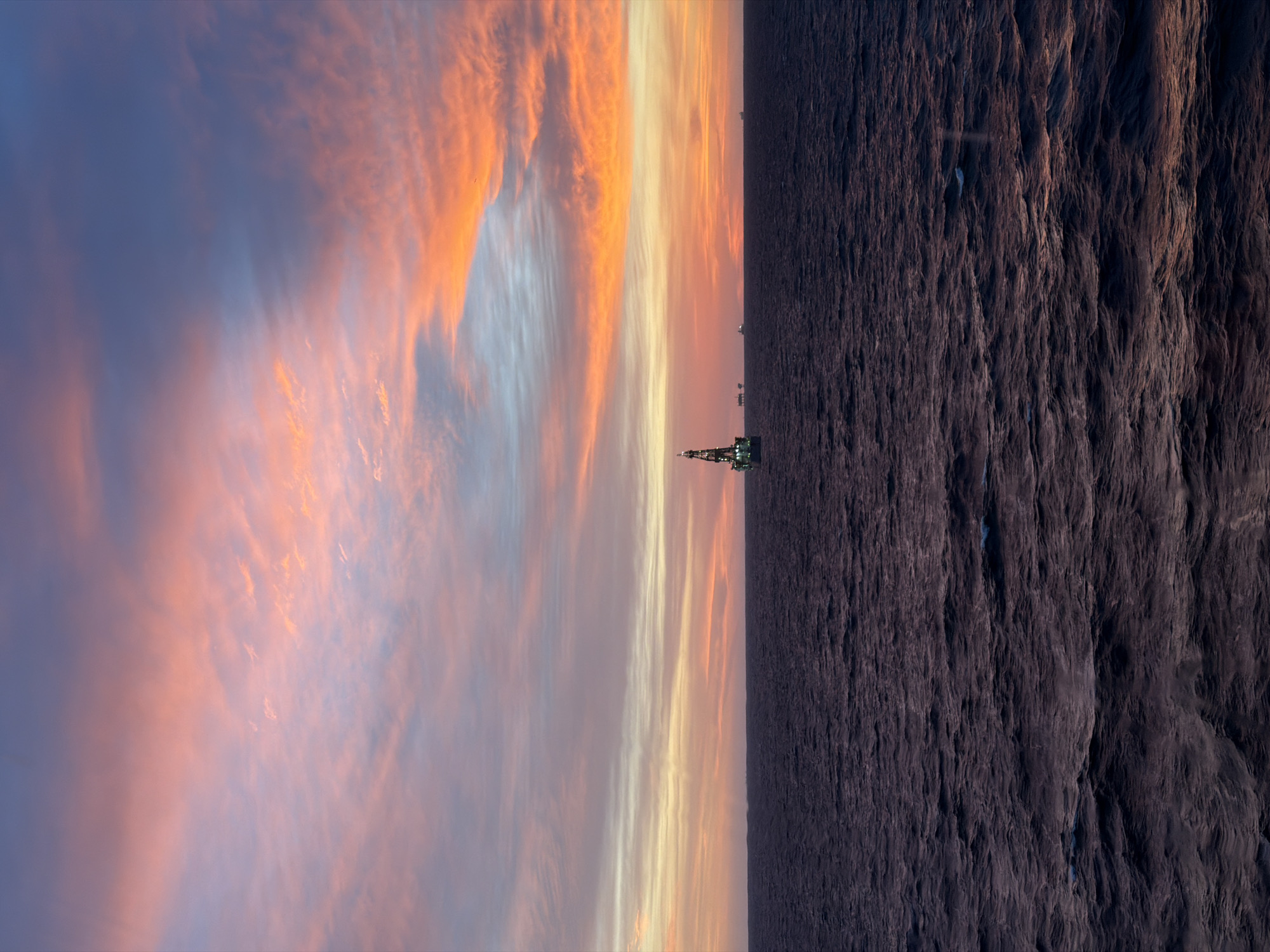 The image, when viewed in landscape orientation (rotated 90 degrees clockwise), captures a dramatic sunset or sunrise over a vast body of water, likely an ocean or sea. The sky dominates the upper portion of the frame, painted with striking horizontal streaks of vibrant orange, yellow, pink, and softer blue clouds, indicating the low angle of the sun. The colors transition from warm, intense hues near the horizon to cooler, more subdued blues and purples higher up.
On the horizon, silhouetted against the colorful sky, stands a prominent offshore structure, possibly an oil rig or drilling platform, which is partially illuminated by green lights. Several other smaller, distant structures or vessels can be faintly seen along the dark horizon line.
The lower half of the image is comprised of the dark, rippling surface of the water, which appears to be in shadow or reflecting the deep blue of the sky. The water's texture is uneven and turbulent, with visible waves and currents creating a complex pattern of light and shadow on its surface.
There is no discernible human activity depicted, and no visible text in the image. The scene conveys a sense of grandeur and isolation, characteristic of open sea environments at dawn or dusk.