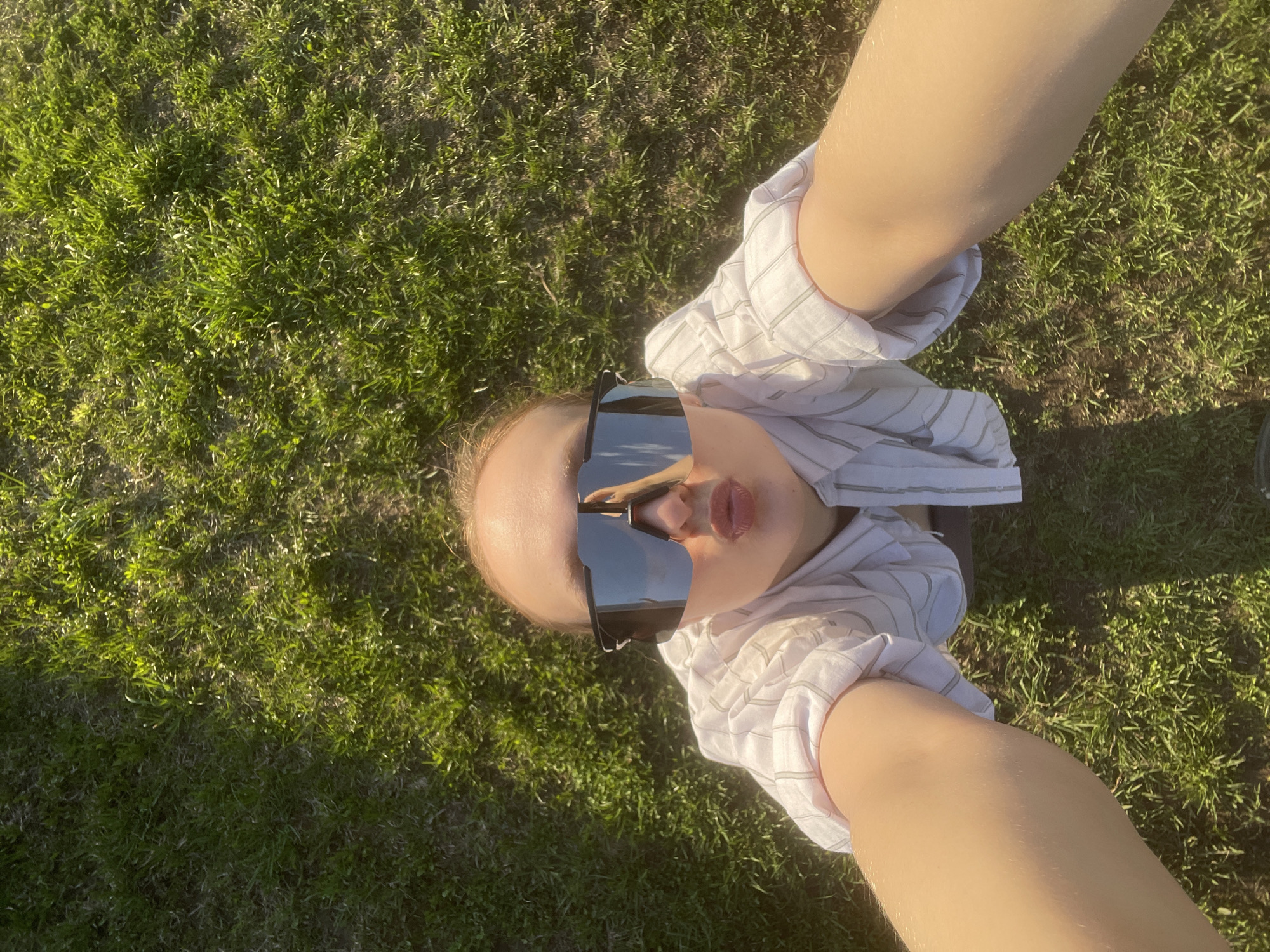 A young person is pictured from a low-angle perspective, lying on a patch of green grass. They are wearing a striped white shirt and large, dark sunglasses that reflect the environment. The location is Подшивалово, Russia. The person appears to be taking a selfie, as their arm is visible extending towards the camera. The background is primarily green grass, indicating an outdoor setting such as a park or yard.