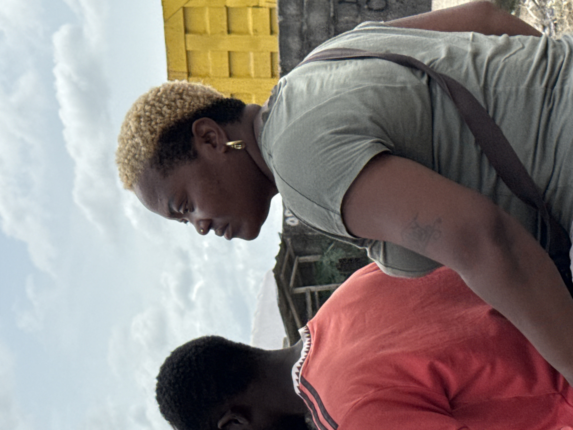 This outdoor image captures two individuals, likely young adults, from a slightly elevated and tilted perspective, set against a cloudy sky and urban background.
The primary subject, positioned in the upper portion of the frame, is a person with dark skin and short, curly, bleached blonde hair, with dark roots showing. They are wearing a light olive green t-shirt and have a dark brown strap, possibly from a bag, draped across their shoulder and chest. A small gold hoop earring is visible in their right ear, and a tiny nose stud adorns their nose. A faded tattoo can be seen on their right arm. The person is bending forward, looking downwards with an expression of concentration or pensiveness.
Partially visible in the lower portion of the frame is a second individual, also with dark skin, wearing an orange or coral-colored t-shirt that features dark stripes and a white patterned trim around the neckline. This person is also bent forward, implying they are engaged in a similar activity or observing something below the frame.
The background consists of a bright, cloudy sky, indicating daytime. In the upper right corner, a textured, bright yellow structure, possibly part of a building, is visible. Further back, indistinct darker structures or foliage can be discerned. The overall setting suggests an urban or semi-urban environment in Ado, Nigeria. No visible text is present in the image.