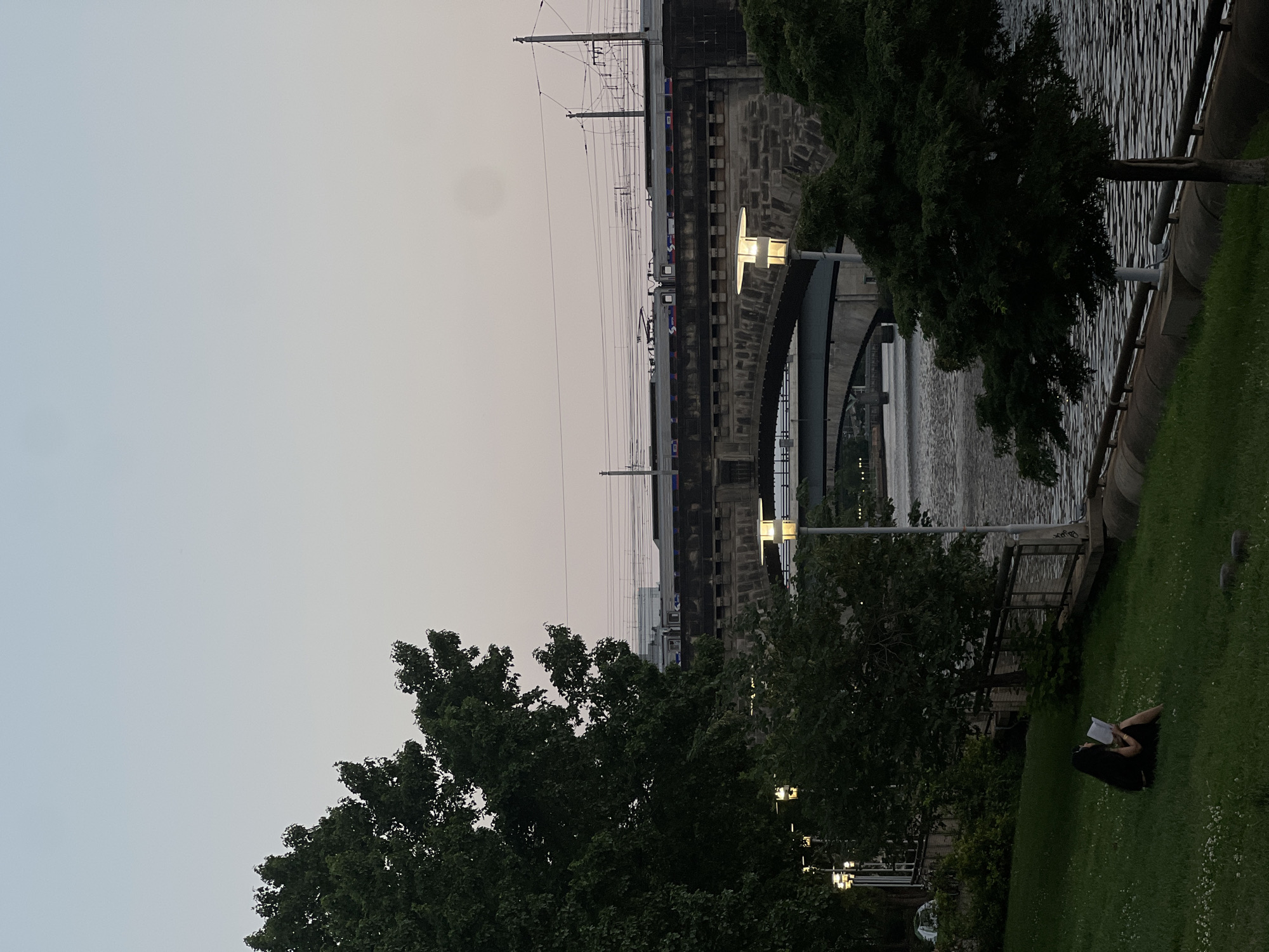 In a park setting in Philadelphia, Pennsylvania, USA, a person sits on a grassy lawn near the Schuylkill River, reading a book. Behind them, a train travels across a stone bridge. The sky is a pale gradient of pink and blue, indicating either early morning or late evening. Trees frame the scene, with the river flowing gently in the mid-ground and a second, larger bridge visible further back. Streetlights are illuminated along the riverbank.