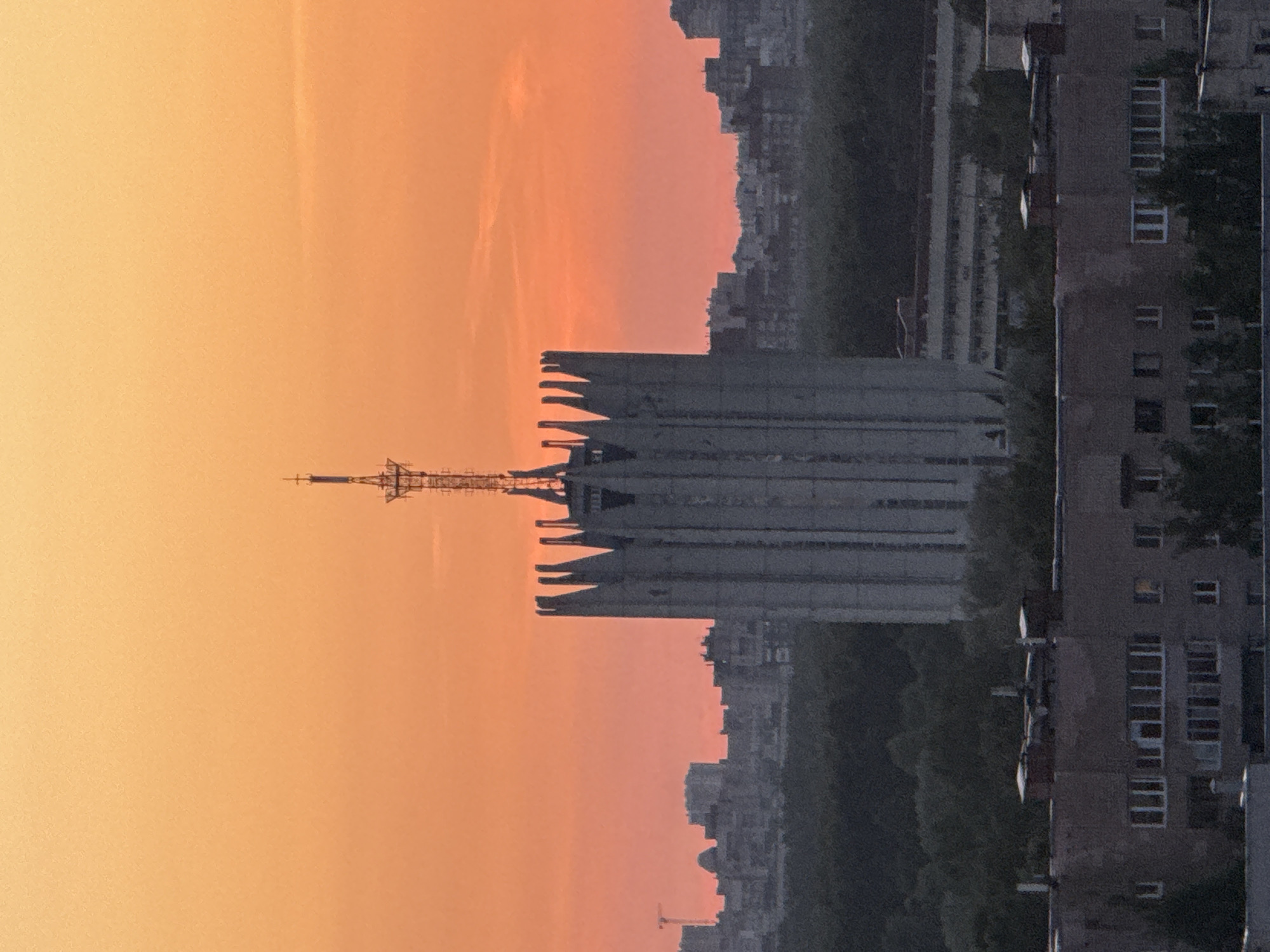 The image displays an urban cityscape at either sunrise or sunset, characterized by a vibrant gradient sky and silhouetted buildings. The sky transitions from a soft yellow-orange at the top to a deeper orange-pink towards the horizon, with faint atmospheric streaks or a contrail visible in the upper portion.
The primary subject is a large, imposing building with distinctive architectural features, including a series of upward-curving, fin-like structures along its roofline. A tall, lattice-structured antenna or mast extends prominently from the center of its roof. This building strongly resembles the Russian Academy of Sciences Presidium in Moscow. To the right of this central structure, and extending further into the frame, are several multi-story residential or administrative buildings, appearing dark and monolithic against the brighter sky. These buildings are typical of post-Soviet architecture, with visible window patterns.
In the mid-ground and background, additional urban structures are visible, forming a dense skyline, largely obscured by the low light conditions and appearing as dark masses. Patches of dark green foliage, indicative of trees, can be seen interspersed among the buildings, especially in the areas between the main structures and the distant skyline.
No people are visible, and no specific activities are taking place. The scene conveys a sense of quiet urban expanse during a transitional period of the day. There is no visible text in the image. The overall impression is one of an expansive city, possibly in Russia, captured in the dramatic light of dawn or dusk.
