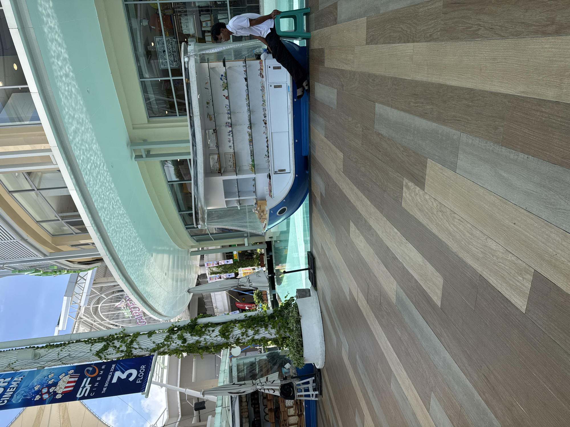 The image shows an outdoor area in Patong, Thailand, with a walkway made of light brown and grey wooden planks. In the background, a modern building with large windows and light-colored facades is visible. A curved, light blue awning extends from the building, casting dappled shadows on the walkway.
In the center of the image, a stall designed to look like a blue and white boat is placed next to a pool of clear blue water. The stall displays various small items on shelves, resembling a souvenir shop. A young man wearing a white polo shirt and dark pants is sitting on a green plastic stool to the right of the boat stall, facing the stall and looking down. He appears to be resting or attending the stall.
To the left of the boat stall, another smaller stall with an umbrella is visible, displaying baskets of goods. A white stool and a speaker are placed next to this stall. A vertical pole with climbing green vines is situated between the two stalls, with a blue banner advertising "SFC CINEMA" and "THE BOTANICA ZONE 3 FLOOR" attached to it. The environment suggests a commercial or recreational area, possibly a shopping mall or promenade in Patong. The sky above is bright blue with some clouds.