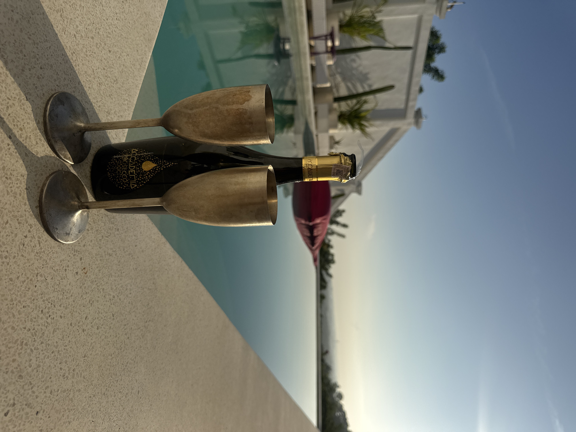 The image depicts a bottle of Accademia Prosecco DOC wine laid horizontally on a concrete ledge with two metal champagne glasses positioned on either side. The location appears to be in Kuta Selatan, Indonesia, near a pool. In the background, a white building with plants and a clear blue sky are visible.