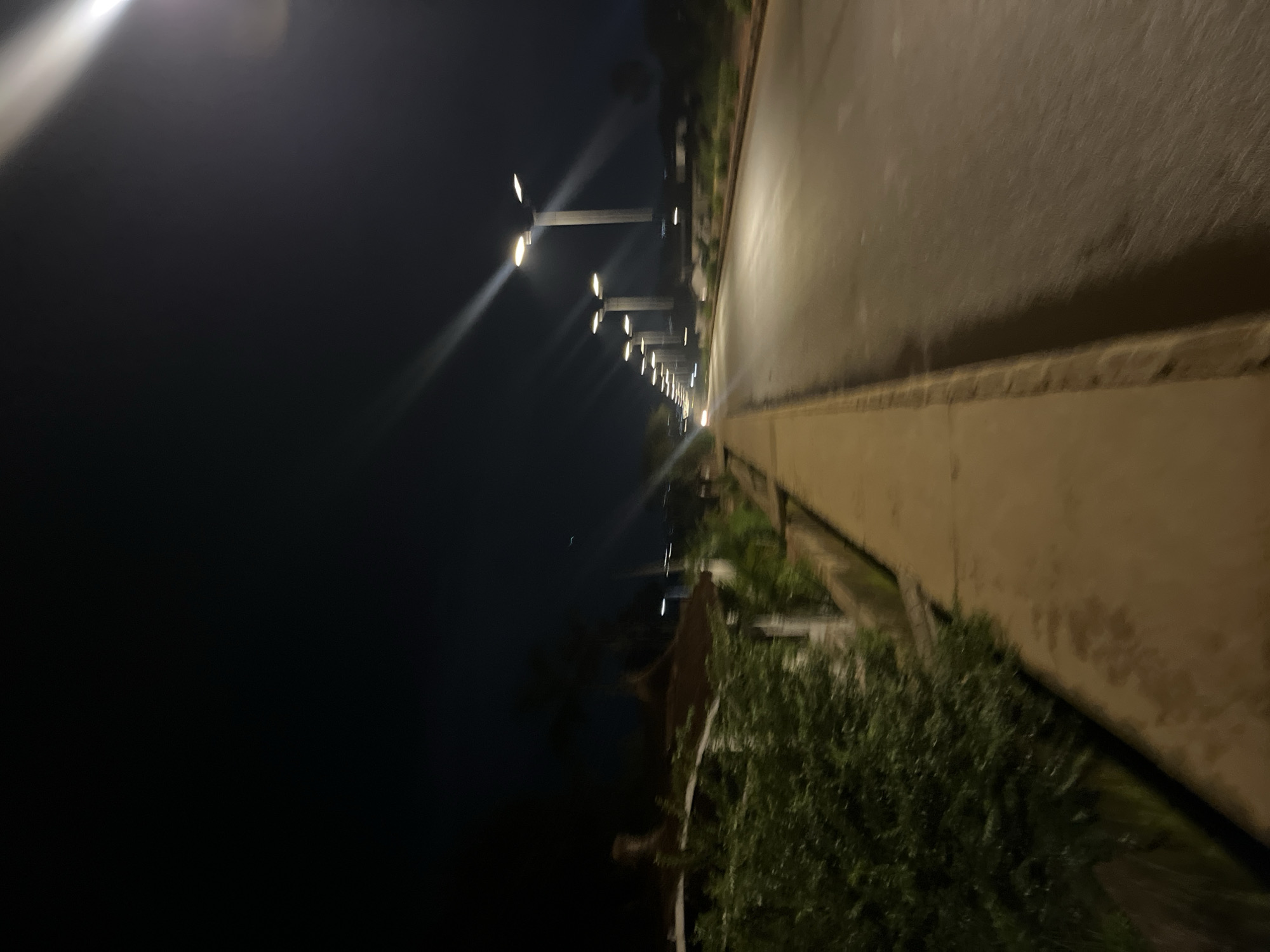 The image captures a street scene at night in Bukuru, Nigeria, illuminated by streetlights. A paved road stretches into the distance, lined on the left by a concrete sidewalk and lush green vegetation. A row of modern streetlights with multiple lamps casts bright beams of light across the road and sidewalk, creating stark shadows and lens flares. The dark night sky is visible above, with no stars discernible. The overall impression is one of a well-lit urban thoroughfare.