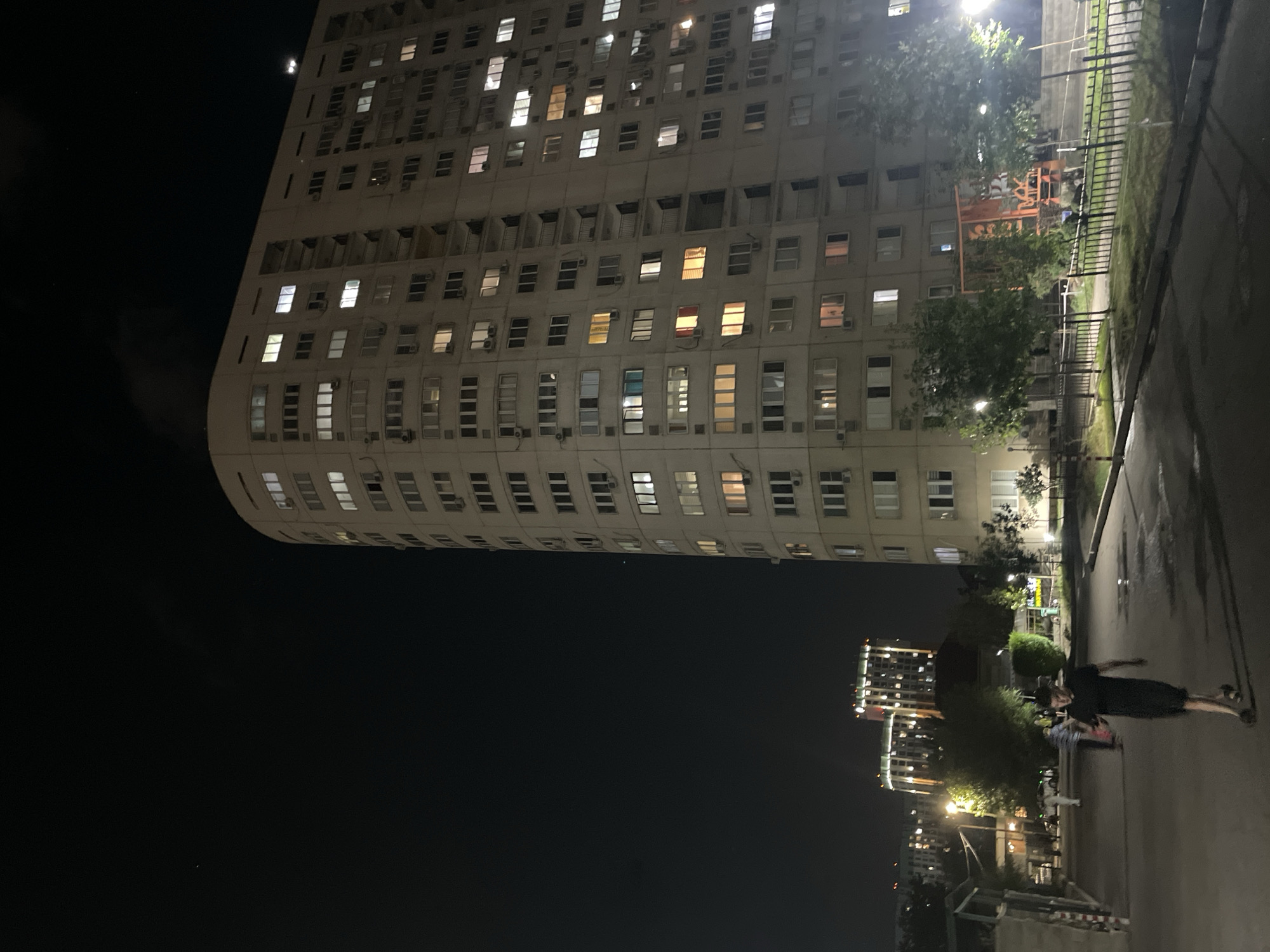 The image captures an urban scene at night in Almaty, Kazakhstan. A large, multi-story apartment building dominates the right side of the frame, its facade lined with numerous windows, many of which are illuminated with warm yellow and white light, suggesting occupied apartments. The building's curved structure is visible against the dark night sky. In the background, to the left, two other apartment buildings are also illuminated, indicating a densely populated residential area.
In the foreground, a paved pathway or street is visible, with a few trees and streetlights lining its edge. A lone male figure, wearing a dark t-shirt and shorts, is walking along the pathway away from the camera, his shadow cast on the ground. Several other indistinct figures and parked vehicles are present further down the path, suggesting some activity in the area. The overall impression is one of a typical residential neighborhood in Almaty during the evening. The sky is dark, with a few faint stars or distant lights visible. The architectural style of the buildings suggests a post-Soviet era design, common in many Central Asian cities.
