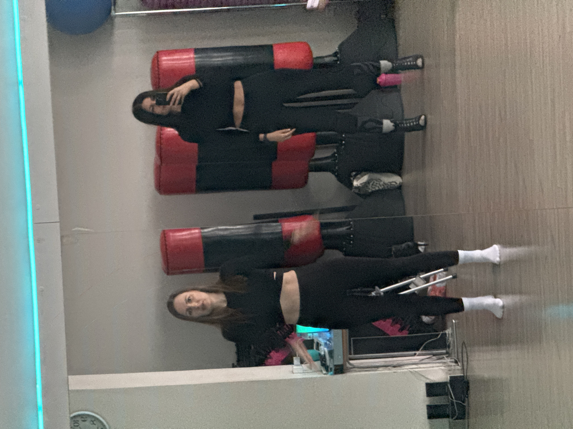 A person stands facing a mirror in what appears to be a gym, taking a selfie with a mobile phone. The person is wearing black athletic clothing, with bare midriff. Her image and reflection are visible. In the background, one can see red and black cylindrical objects, presumably part of gym equipment. A blue exercise ball is visible in the upper left. The environment consists of mirrored walls, gym equipment and a light colored floor. It is impossible to determine the location in the city of Chaikovsky, Russia, based on the available information.