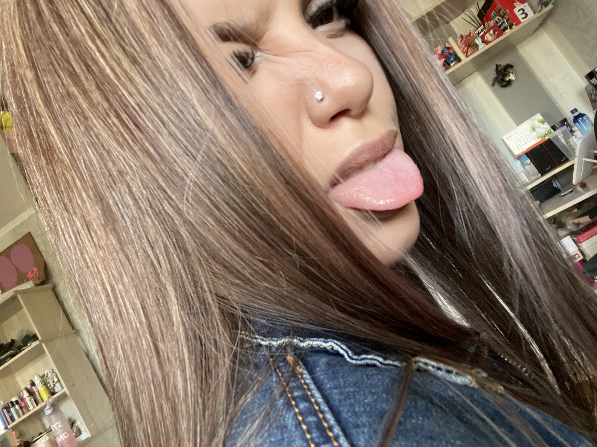 The image is a close-up, low-angle, selfie-style photograph of a young woman sticking out her tongue. She has long, straight, light brown hair that partially covers her face. She is wearing a blue denim jacket. A small, diamond stud is visible in her nostril. Her lips are covered in a matte, nude-colored lipstick.
In the background, there are shelves filled with various items. On the left shelf, there are rows of small bottles, possibly cosmetics or nail polish. Further back, there's a wooden organizer with compartments. On the right, a shelf displays an assortment of colorful items, including what appear to be craft supplies or small decorative objects, and a calendar with a picture of flowers. The overall environment suggests an indoor setting, possibly a bedroom or dressing area. The lighting is soft and natural. There is no indication of the specific location within the city of Vsevolozhsk, Russia, or any environmental context that would specify the city.