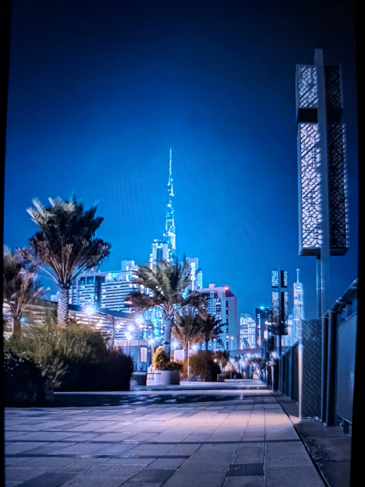 The image depicts a nighttime cityscape with prominent illuminated skyscrapers against a deep blue sky. In the foreground, a paved walkway with geometric patterns is visible. To the left, several palm trees stand silhouetted against the glowing buildings. The background is dominated by a cluster of modern architecture, with the most striking structure being a very tall, slender skyscraper adorned with lights. On the right side of the frame, a tall, modern structure with a patterned facade is partially visible. The scene is urban and suggests a well-lit, developed area. The location provided, Yamunanagar, India, does not align with the architectural style of the iconic Burj Khalifa-like skyscraper visible in the image, which is characteristic of Dubai. Therefore, it is unlikely that this image was taken in Yamunanagar, India.