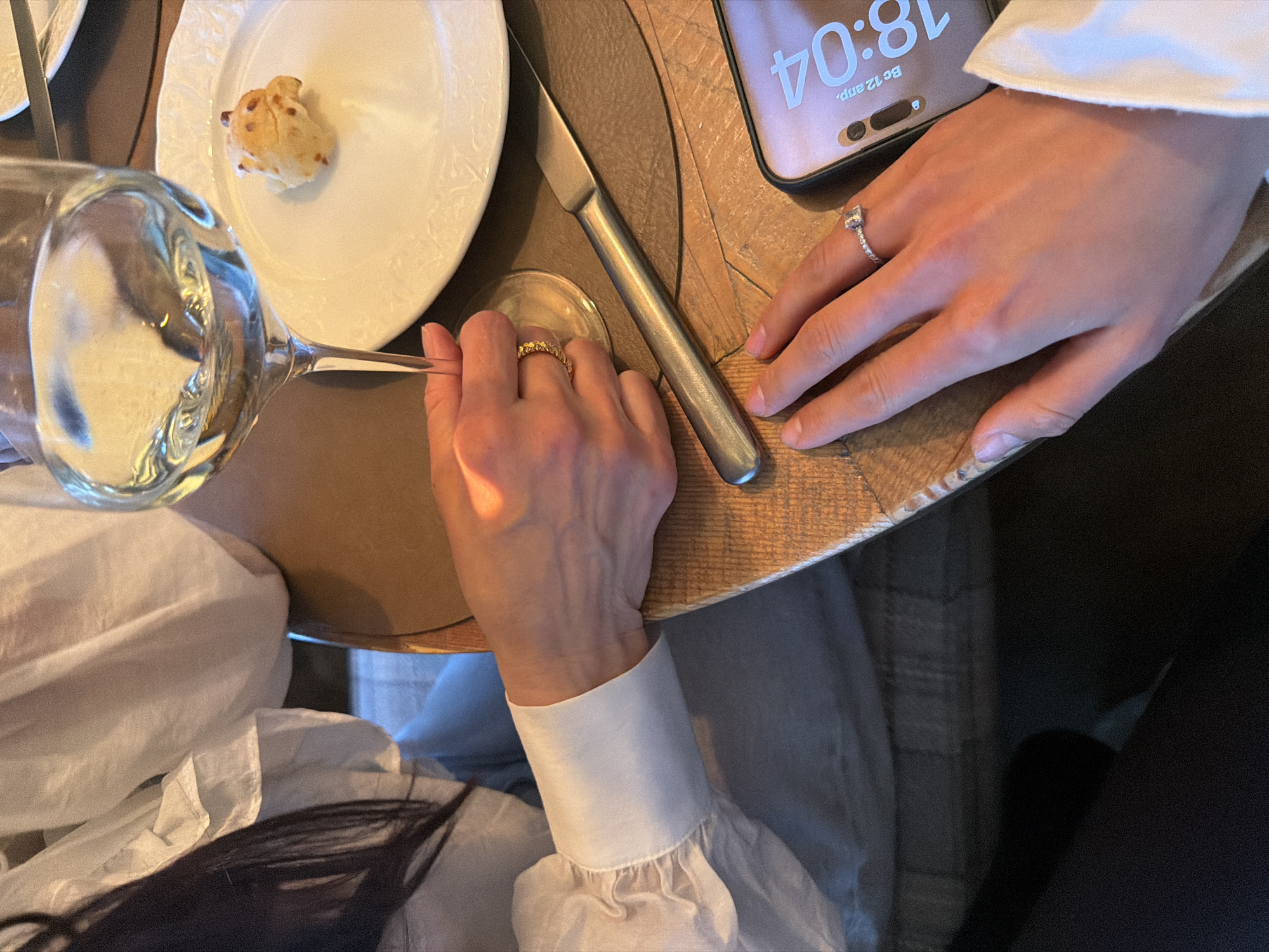The image shows an overhead view of a dining table scene. A person wearing a white blouse with long sleeves is seated at the table. On the table, there is a white plate with a small bread roll, a glass of water, a knife, and a smartphone. The smartphone screen displays "18:04." One hand has a ring on the finger. The location appears to be a restaurant setting. There is no definitive information in the image to confirm it is Krasnoyarsk, Russia.