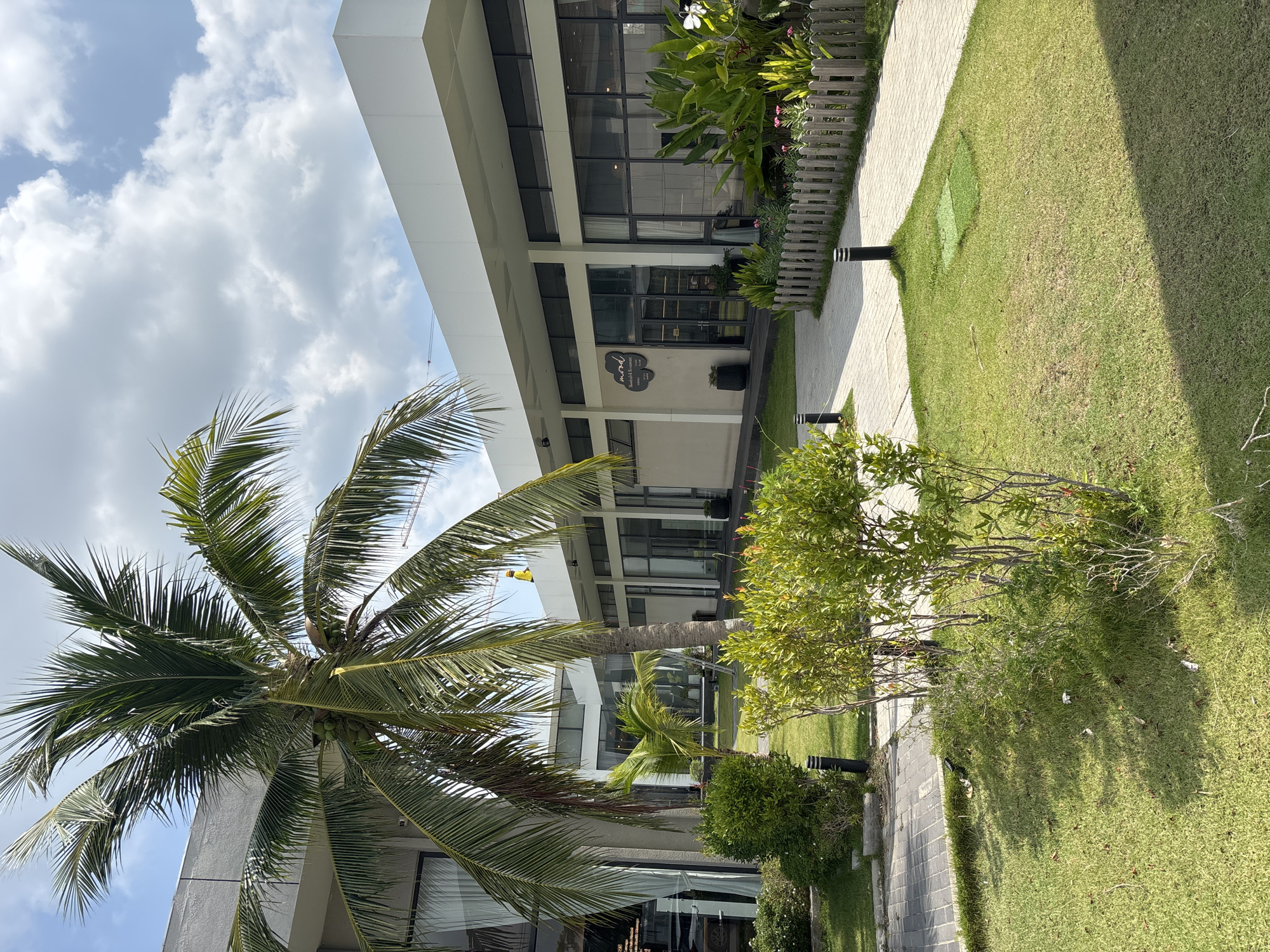 This image captures a vibrant, sunlit outdoor scene in Choeng Thale, Thailand, featuring a modern building surrounded by lush greenery. The time of day appears to be bright daytime, with clear skies interspersed with fluffy white clouds, casting distinct shadows on the ground.
A prominent feature is a contemporary building, likely a resort, restaurant, or commercial establishment, characterized by its clean white and gray architectural lines and large, dark-paned windows reflecting the surroundings. A black sign on the building clearly displays the name "mood" in a stylized script, followed by "kitchen & bar" and smaller text indicating services like "breakfast," "lunch," "dinner," and "coffee," along with approximate operating hours.
In the foreground and mid-ground, several large palm trees with coconuts are visible, one particularly dominating the left side with its fronds fanning out against the sky. Various smaller shrubs and leafy plants with some floral accents line the building's facade and pathways. A neatly manicured green grass lawn extends across the right side of the frame, bordered by a light-colored paved walkway, possibly concrete, that curves through the landscape. Another gray-tiled path is visible in the lower right corner. Black, cylindrical outdoor lighting fixtures are placed along the walkways. A decorative, low wooden fence separates some of the planted areas. In the distant background, a construction crane is partially visible against the sky, suggesting ongoing development in the area. The overall impression is one of a well-maintained and inviting tropical setting.