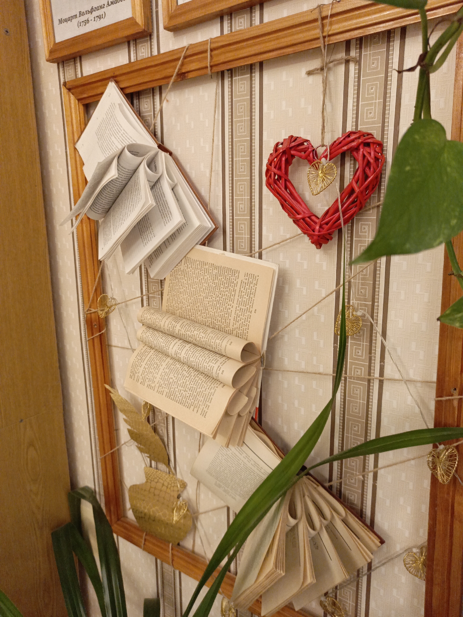 The image displays an indoor decorative arrangement on a patterned wallpapered wall in Markovskii, Russia. The central feature is a rectangular wooden frame from which a creative display is suspended using twine. No people are visible in the scene.
The subjects of the display include several old books, whose pages have been artfully folded, rolled, and fanned out, giving them a sculptural quality. These books are attached to the twine and positioned within the frame. A prominent red wicker heart, with a smaller golden wire heart nested inside it, hangs from the top center of the frame. Several other small golden wire hearts are strategically placed along the twine that crisscrosses the frame. Lush green leaves from a plant are visible in the foreground and intertwining with the display, adding a natural element to the composition.
Above the main frame, part of another wooden frame is visible, containing text in Cyrillic. This text identifies "Моцарт Вольфганг Амадей" (Mozart Wolfgang Amadeus) and his lifespan "(1756-1791)". The open pages of the old books also show various snippets of Russian text, including words like "Сударь", "смерти буду молиться", "чувство", "говорите", and "бог", along with page numbers such as "220", "194", and "239".
The scene suggests a cozy or culturally themed interior, possibly a home, library, or café. The arrangement blends literary elements (books), natural motifs (plant leaves), and romantic symbols (hearts), creating a unique and aesthetically pleasing wall decoration. The lighting is bright and even, indicating either daytime or a well-lit indoor environment.