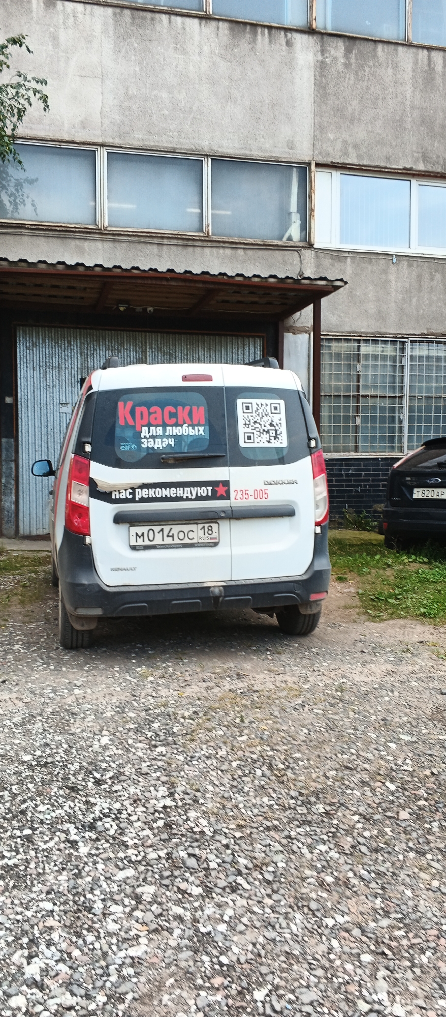 This image depicts the rear of a white Dacia Dokker van parked on a gravel lot in front of a building in Izhevsk, Russia. The van has several stickers on its rear door. The largest sticker, in red and white, reads "Краски для любых задач" which translates to "Paints for any task." To the right of this sticker is a QR code. Below the "Paints for any task" sticker, another sticker reads "нас рекомендуют" (they recommend us) with a red star and the number "235-005". The van's license plate is visible and reads "M014OC 18". The building behind the van is constructed of concrete and has several long, horizontal windows. To the right of the van, a portion of a black car is visible. The ground is covered with gravel and some sparse vegetation.