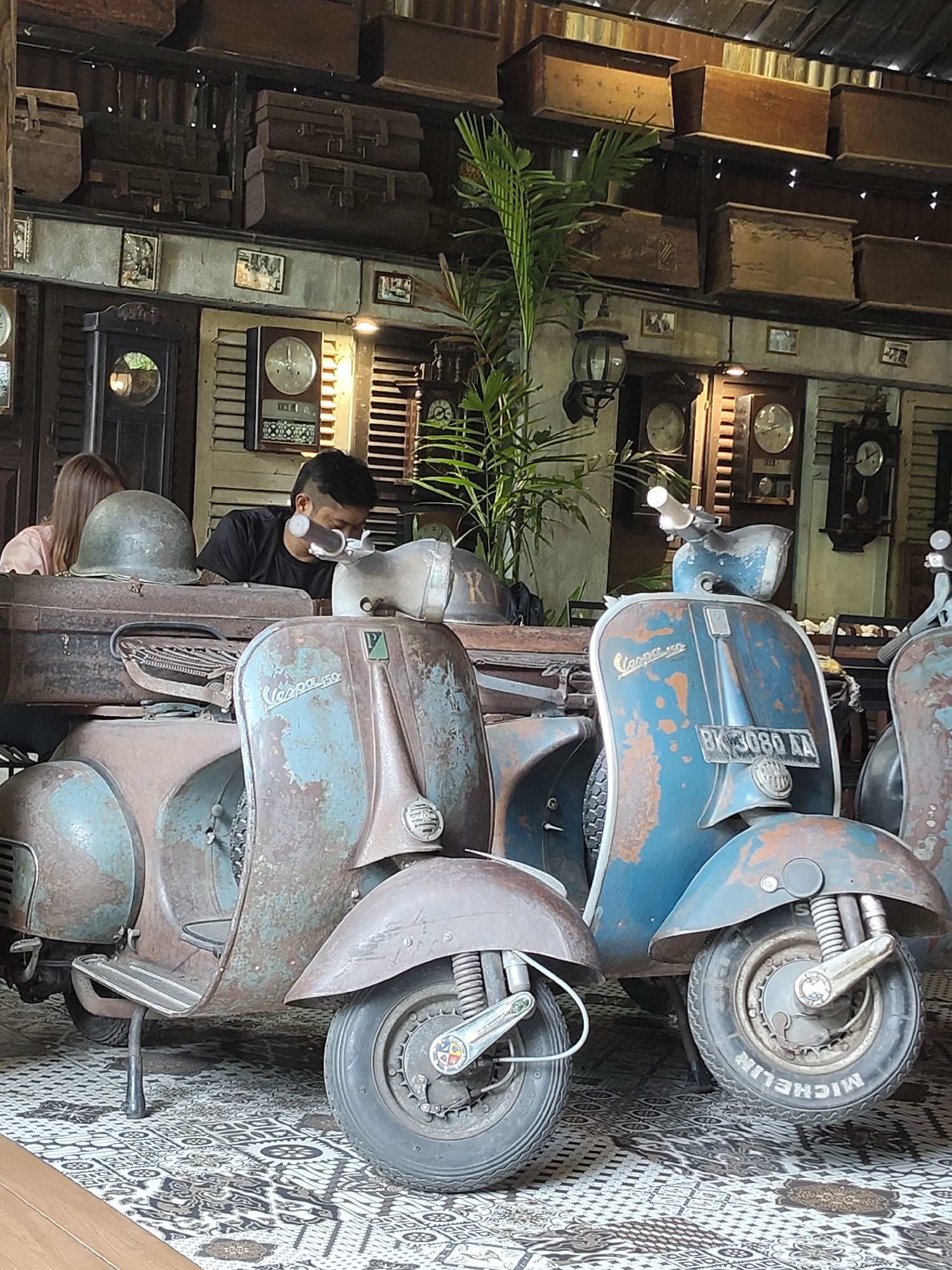 A medium shot captures two vintage Vespas parked indoors, in a setting reminiscent of a retro-themed cafe or shop in the City of Medan, Indonesia. The Vespas, one predominantly rusted brown with blue accents and the other a faded blue with rust, are positioned close together on a patterned floor. A man wearing a black t-shirt and a face mask is seated on the brown Vespa, looking down. To his left, a person with long brown hair, wearing a light pink top, is partially visible. The background features a wall decorated with antique clocks, wooden chests stacked on shelves, and what appears to be a slatted wooden door. A lush green plant stands behind the Vespas, adding a touch of life to the vintage scene. The overall atmosphere is one of rustic charm and nostalgic decor.