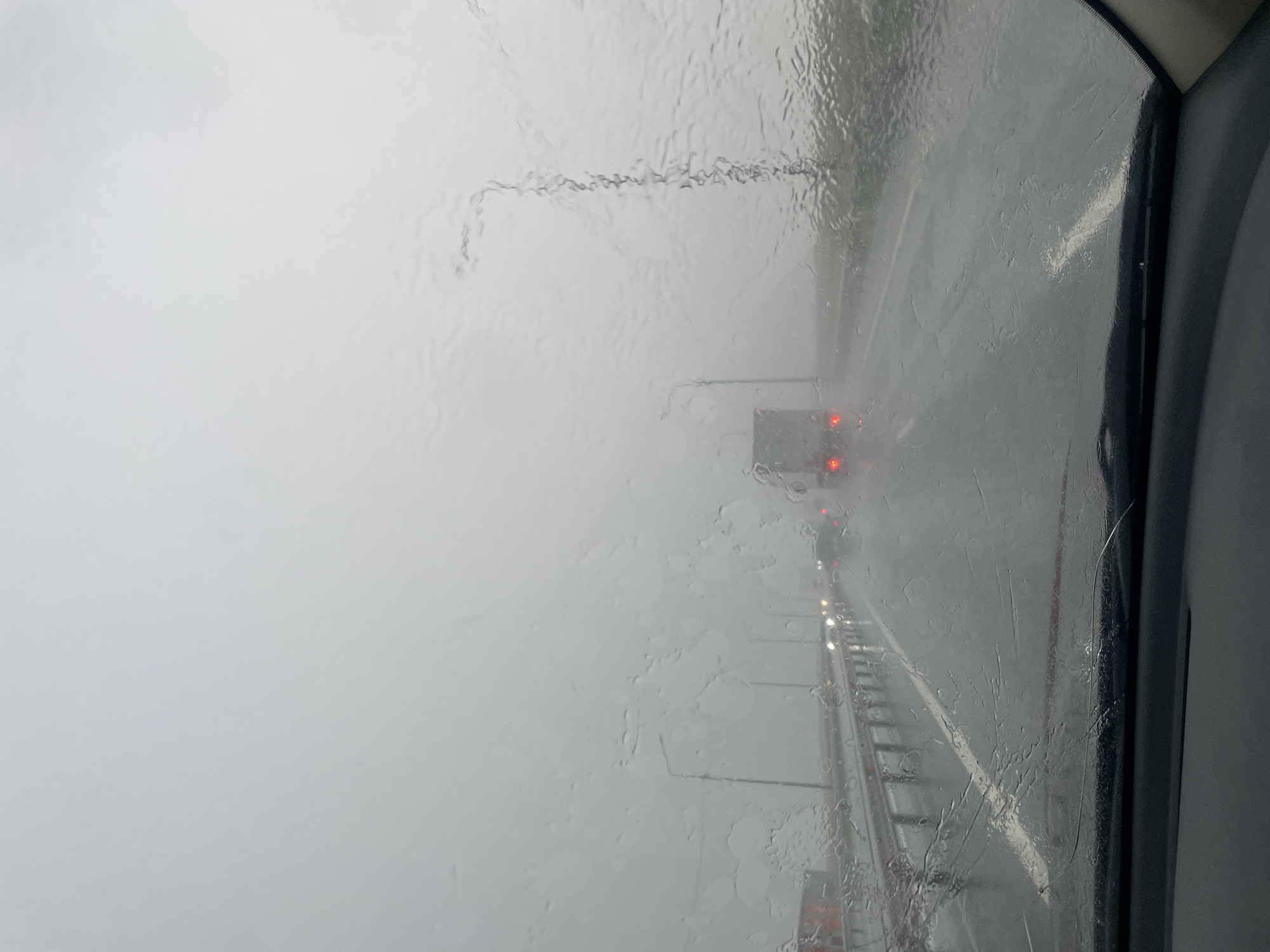The image depicts a view through a rain-streaked windshield of a vehicle traveling on a highway. The weather is overcast and foggy, with heavy rain obscuring visibility. Multiple vehicles, including trucks and cars, are visible on the multi-lane road, their headlights and taillights illuminated. Roadside infrastructure, such as guardrails and streetlights, can be seen through the fog and rain. The road surface appears wet and reflects the lights of the vehicles. The interior of the vehicle, including the dashboard, is visible at the bottom of the frame. The scene suggests a challenging driving condition due to inclement weather.