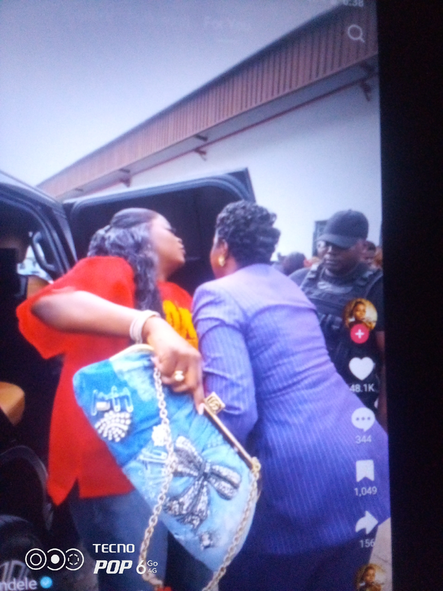 The image depicts a scene with three individuals, two women and a security personnel, positioned in what appears to be an outdoor urban setting in Howayi, Nigeria. The focus is on the interaction between the two women. The woman on the left, wearing a bright red t-shirt with yellow lettering, is looking towards the woman on the right. She is holding a blue purse adorned with intricate designs, including a large bow and sparkling embellishments. Her left arm is extended, and she is wearing bracelets and a ring.
The woman on the right is dressed in a purple pinstriped garment and faces away from the camera, appearing to be engaged in a conversation with the first woman. Her hair is styled in an updo. Behind them, a security guard in a dark uniform and cap is visible, looking towards the left. The background suggests an open area with a building structure that has a corrugated roof and what looks like a canopy or awning. The lighting indicates it might be daytime, possibly overcast.
On the right side of the frame, overlaid social media interface elements are visible, including a profile picture in a circle, a heart icon with "48.1K" below it, a speech bubble icon with "344" next to it, a bookmark icon with "1,049" below it, and an arrow icon with "156" at the bottom. Additionally, there is text overlaying the bottom left corner of the image that reads "TECNO POP 6 Go 4G" and a partial word "ndele" at the very bottom left. The overall mood seems to be one of interaction and perhaps a public event or gathering, given the presence of security personnel.