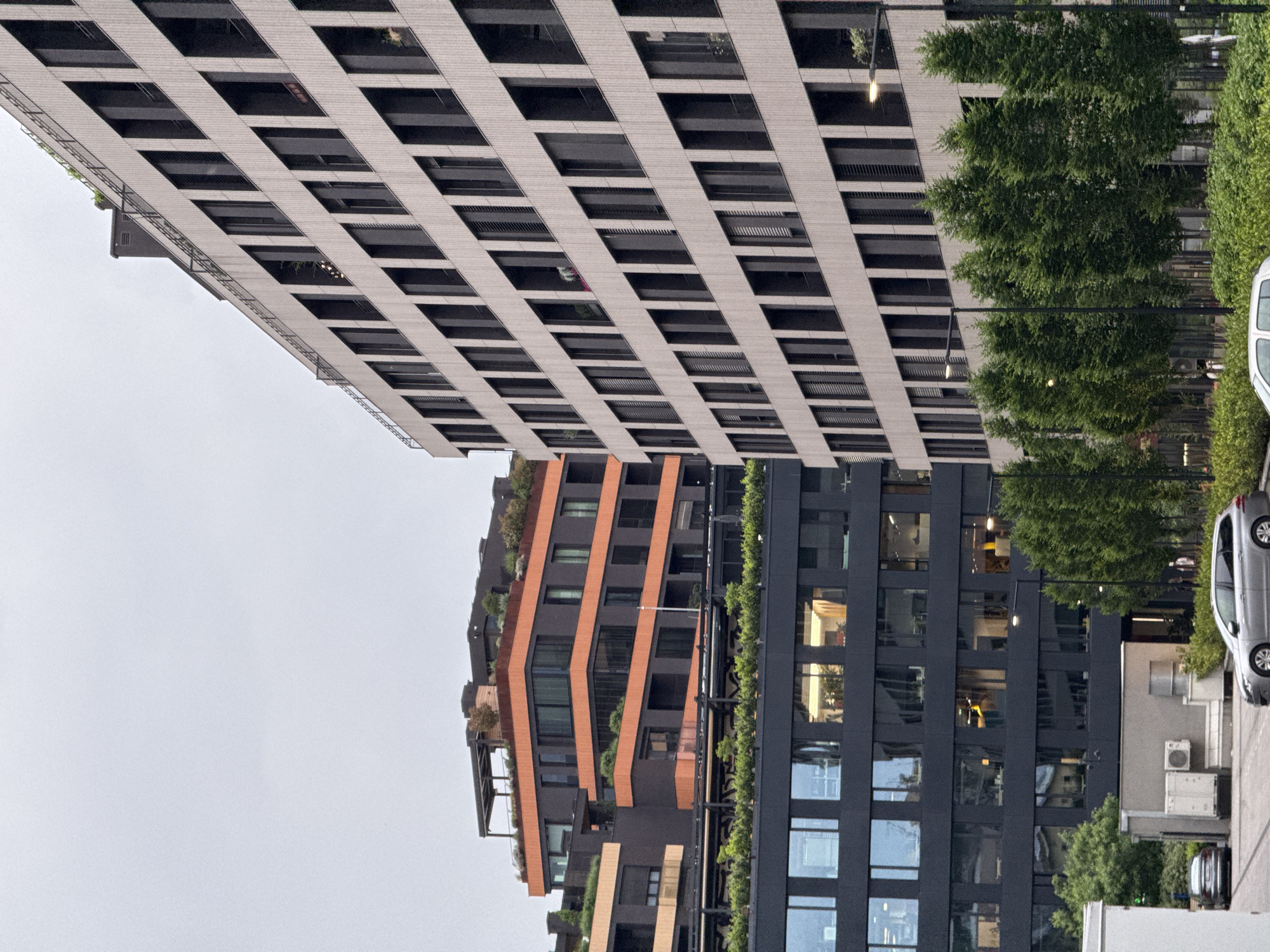 The image shows a street scene in Belgrade, Serbia. There are three buildings visible. The tallest building has a gray and black striped facade with multiple recessed windows. To the right of this building are trees and streetlights. To the right of the trees is a silver car. The second building has an orange, black, and green facade, with an irregular surface. The third building has a dark facade and square windows, with plants growing along its side.