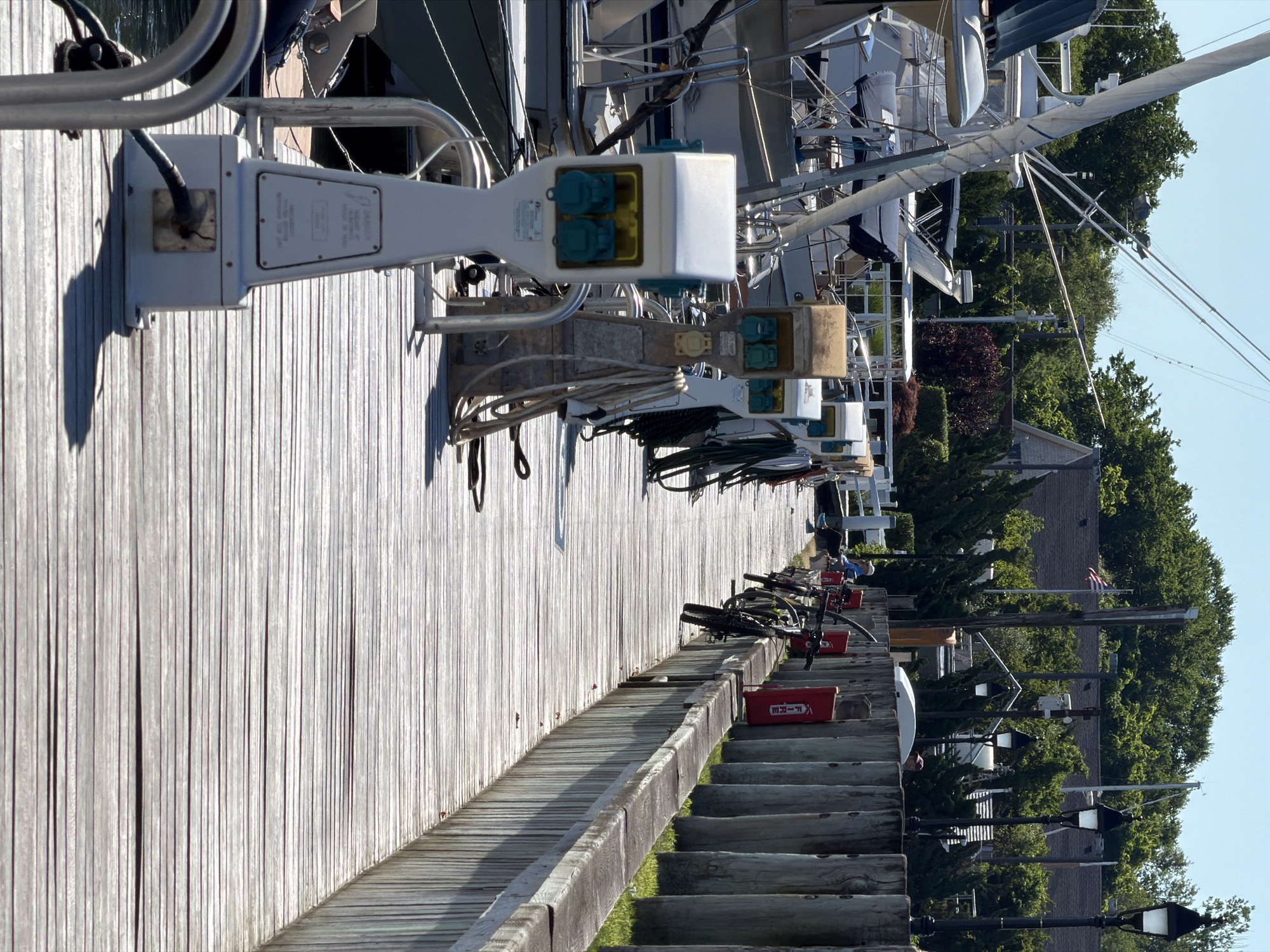 The image depicts a sunny day on a wooden dock in the Village of Sag Harbor, United States of America. The dock is lined with power pedestals for boats, some with coiled ropes. To the left, several sailboats are docked, with their masts and rigging visible against a clear blue sky. Further down the dock, a row of bicycles are parked, with one red fire extinguisher box visible. In the background, lush green trees and houses are seen, with an American flag flying from a flagpole. A few people are present on the dock, including one sitting down and another standing near the bicycles. The scene conveys a sense of a well-maintained marina on a bright summer day.