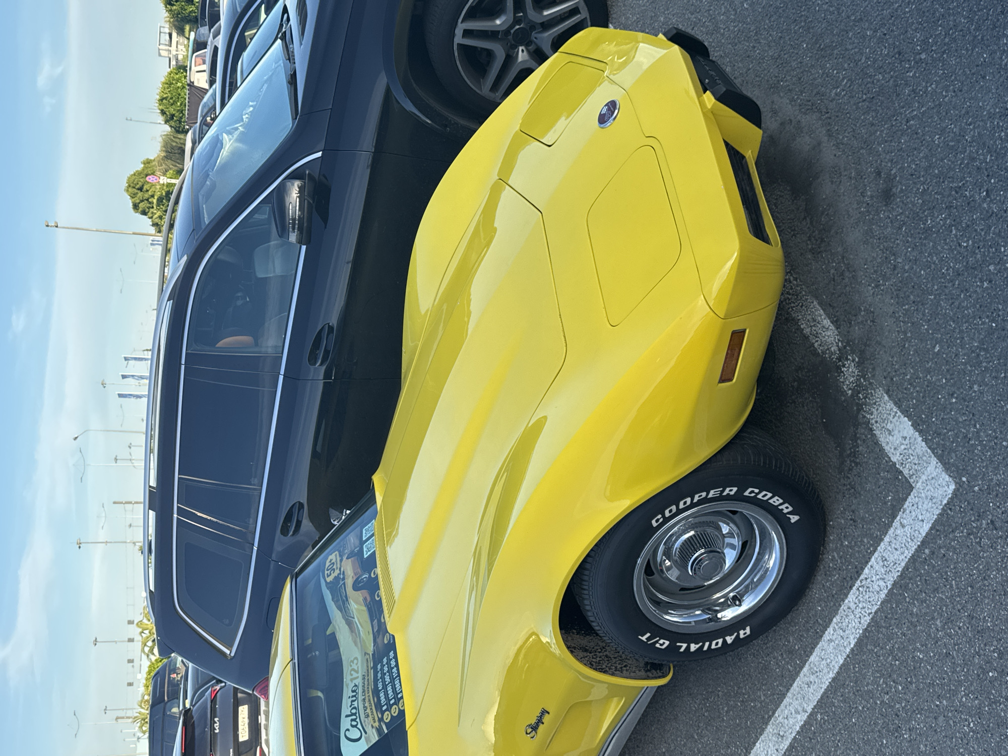 In Sirius, Russia, a yellow Chevrolet Corvette Stingray is parked in a lot, its vibrant color contrasting with the dark SUV beside it. The Corvette's hood prominently displays the "Stingray" emblem, and its front wheel, with a "Cooper Cobra" tire, is visible. A sticker on the windshield reads "Cabrio 123" and features phone numbers. The parking lot surface is asphalt, marked with white lines. The sky above is a clear blue, with a few wispy clouds. Streetlights and trees are visible in the background, suggesting an outdoor urban or suburban setting. The scene is static, capturing a moment in time with no discernible action.