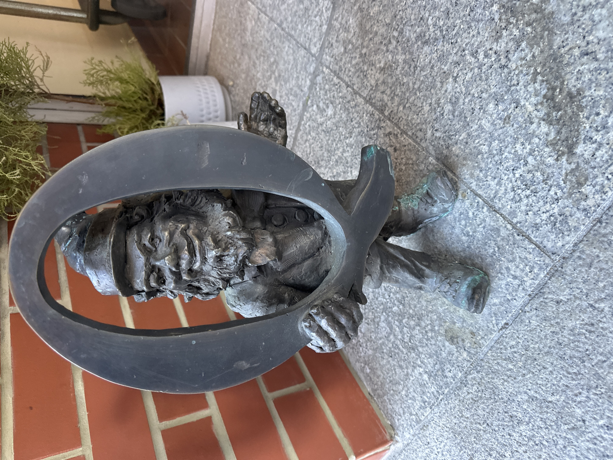 The image shows a bronze dwarf statue, one of the many "krasnale" scattered throughout Wrocław, Poland. This particular dwarf appears to be struggling while trapped inside a large metal fish symbol. The dwarf has a detailed face with a beard and wears a cap. His hands grip the fish symbol. The statue is located on a sidewalk made of granite tiles and is situated near a brick wall and some plants in a white pot.