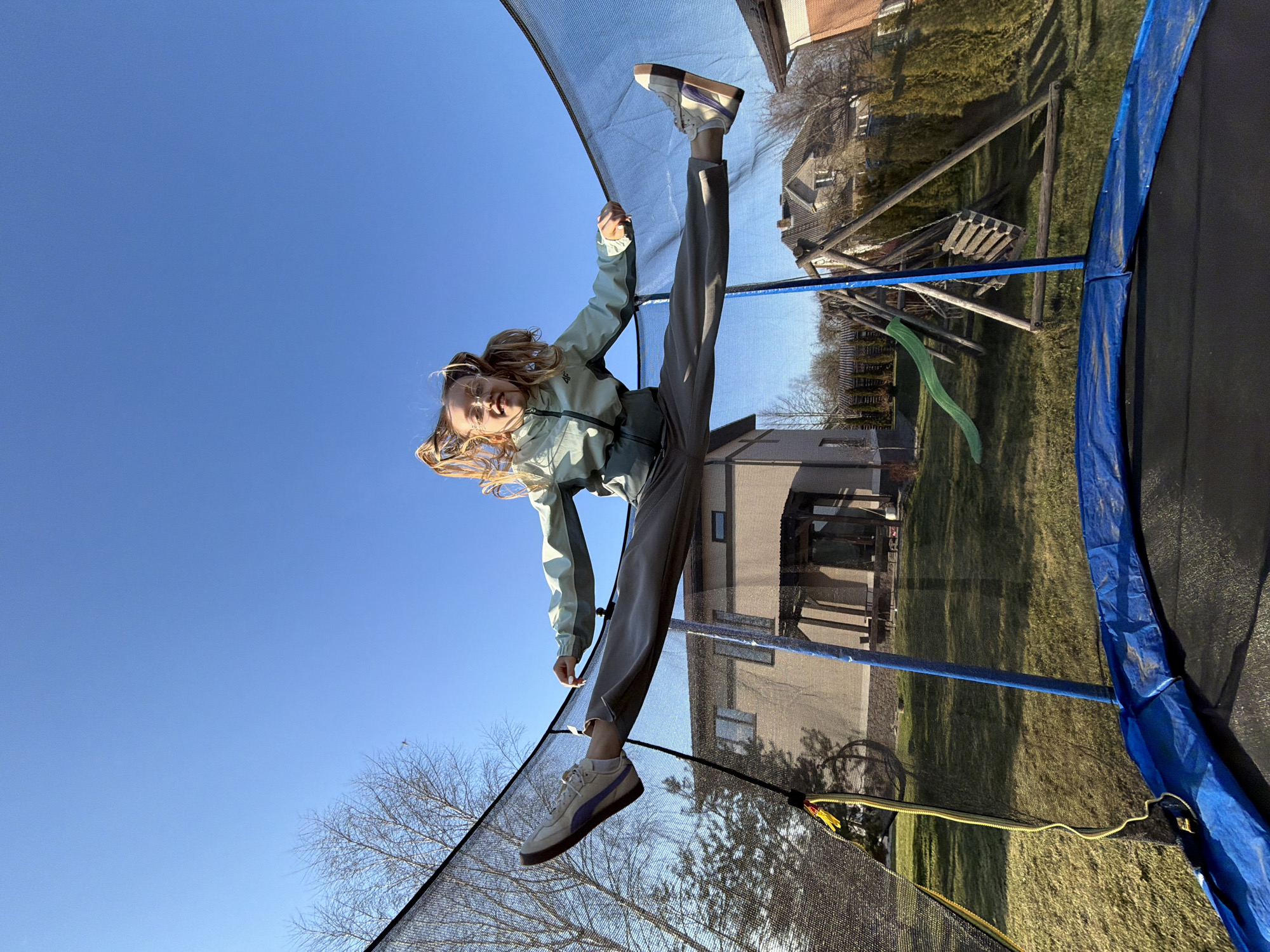 A girl in a light-green jacket and gray pants jumps on a trampoline. The trampoline is surrounded by a black mesh net. The girl is in mid-air, doing a split. Her feet are in sneakers, and her blonde hair is flying. The background includes a clear blue sky, a building with beige siding, a playground with a green slide, and trees without leaves. The location appears to be a backyard in a residential area of Dreiliņi, Latvia.