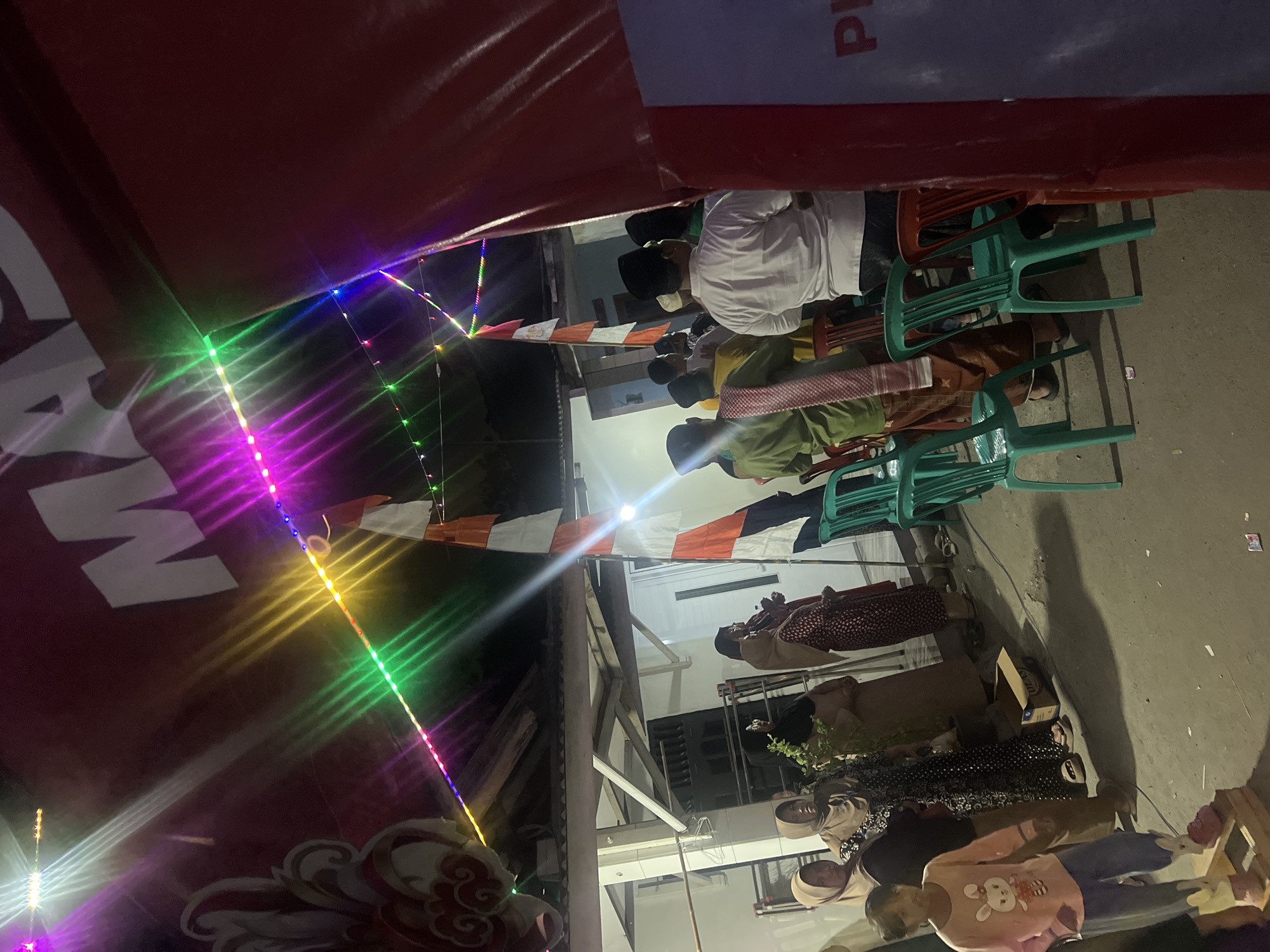 The image captures a nighttime outdoor gathering in Klapanunggal, Indonesia, likely a community event or celebration. The scene is illuminated by colorful LED string lights draped across the top, casting vibrant hues of pink, purple, green, and yellow. In the background, a large red banner with white lettering, partially obscured, suggests a formal occasion.
Several individuals are present, dressed in traditional attire. Women, predominantly wearing hijabs and long dresses, are seen standing and conversing. Men, some in peci caps and shirts, are also present, with some appearing to stand in groups. A young girl in a pink shirt with a rabbit graphic and blue pants stands in the foreground, looking towards the camera.
The setting appears to be a paved outdoor area, possibly a street or courtyard, with a building structure visible behind the attendees, featuring a white wall and roof. Green and red plastic chairs are arranged, indicating seating for guests. A partially opened cardboard box rests on the ground. The ground surface shows scattered debris, consistent with an outdoor event. The overall atmosphere seems social and celebratory, enhanced by the festive lighting.