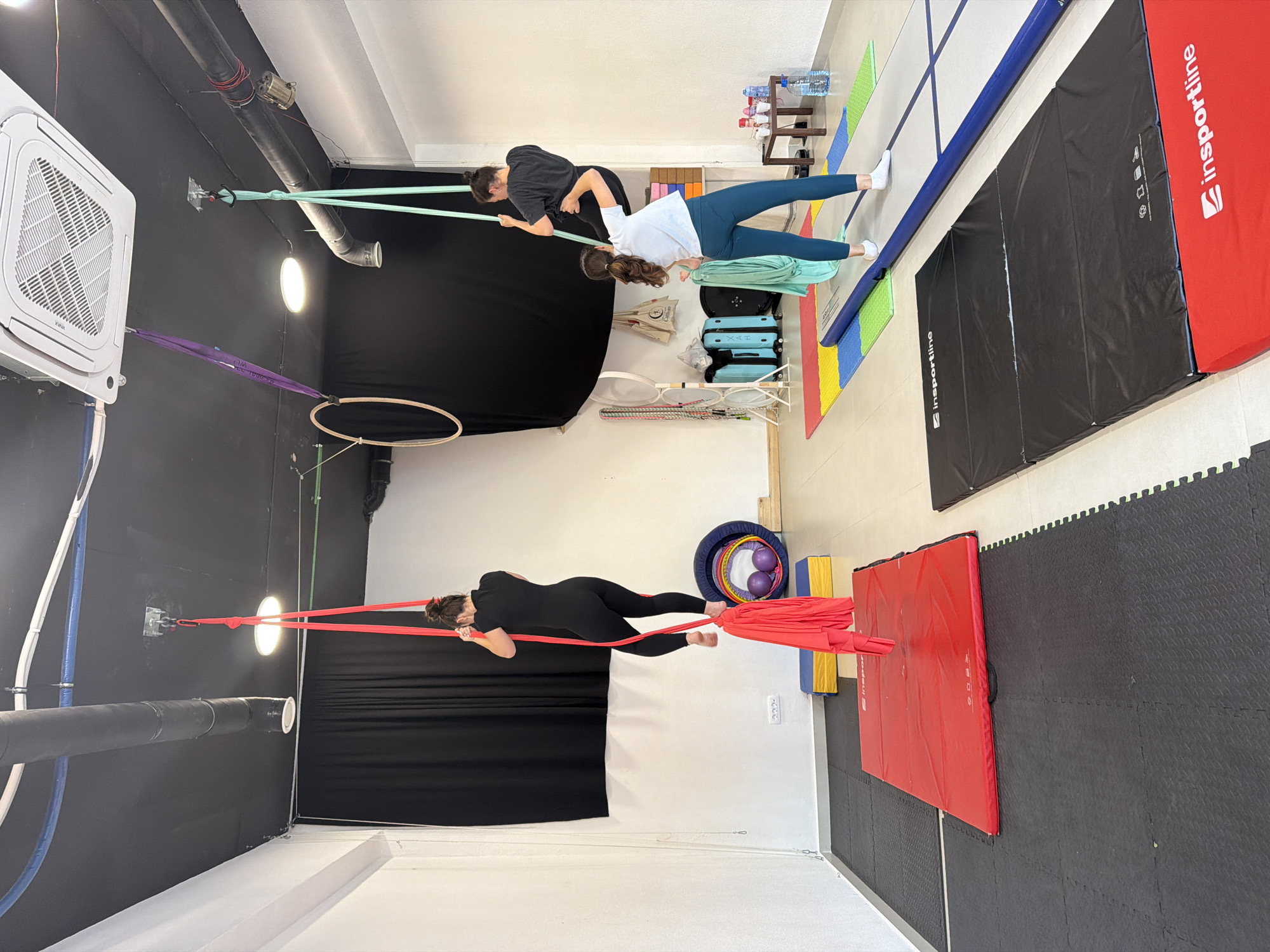 This image captures an indoor fitness or aerial studio in Bar, Montenegro, characterized by its black walls and white ceiling and floor. Two women are actively engaged in exercise.
In the foreground, a woman dressed in black activewear is suspended horizontally by a bright red resistance band attached to the wall. She appears to be performing a core strength or aerial conditioning exercise, her body held taut. Behind her, another woman wearing a white t-shirt and teal leggings stands upright, holding a light green resistance band, seemingly engaged in a resistance training or stretching exercise.
The studio is equipped with various items. Large crash mats, one red and one black, cover parts of the floor for safety. An inflatable blue and grey air track mat is also present. Colorful interlocking foam mats provide additional cushioning. An aerial hoop (lyra) hangs from the ceiling by a purple strap, though not currently in use. Other visible objects include an HVAC unit mounted on the wall, exposed pipes along the ceiling, bright overhead lights, a wooden stand with water bottles, stacked light blue suitcases, and a basket containing hoops and purple balls.
The scene depicts a focused training session. The brand "Insportline" is clearly visible on the red and black crash mats, as well as on the blue air track. The HVAC unit displays the brand "KAVIA." The indoor setting suggests the activity takes place regardless of the time of day or outdoor weather.