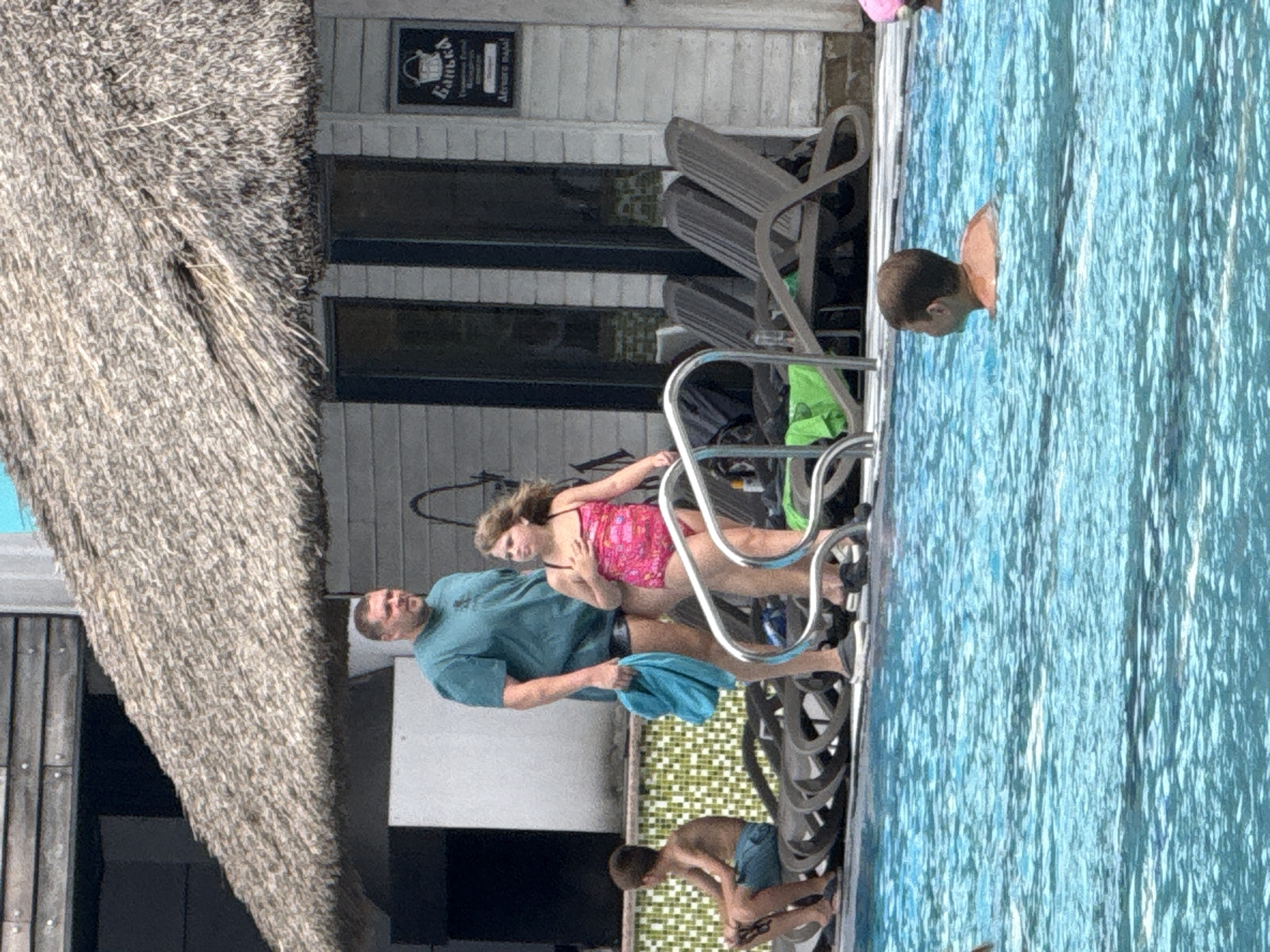 This image shows an outdoor swimming pool area, likely a resort or hotel, situated within the Массандровский поселковый совет, Ukraine. A man in a teal t-shirt and dark shorts stands by the pool, holding a teal towel. Beside him, a young girl in a pink swimsuit with a floral pattern is walking towards the pool. To the left, another boy in blue swim trunks is sitting on a lounge chair. In the background, a thatched-roof structure is visible, with white siding and dark-framed windows. Several lounge chairs are arranged near the building. The pool itself is filled with clear blue water. The overall scene depicts a leisure setting on a sunny day.