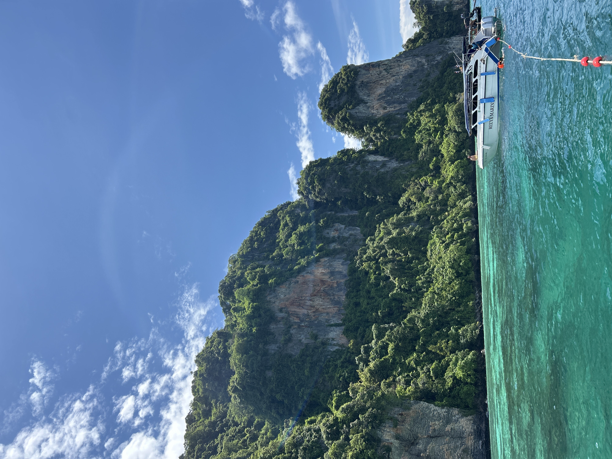 A speedboat with the name "REE YA MARIN" is docked in the turquoise waters off the coast of Ao Nang, Thailand. The boat is white with blue accents and has two engines on the back. Several people are visible on the boat, including one person sitting on the bow and two people near the engines. In the background, lush green cliffs rise from the water, covered in dense vegetation. The sky is a clear blue with scattered white clouds, and the sun is shining brightly. A rope with red buoys is visible in the foreground, connecting to the speedboat. The water is clear and calm, with sunlight reflecting off the surface.