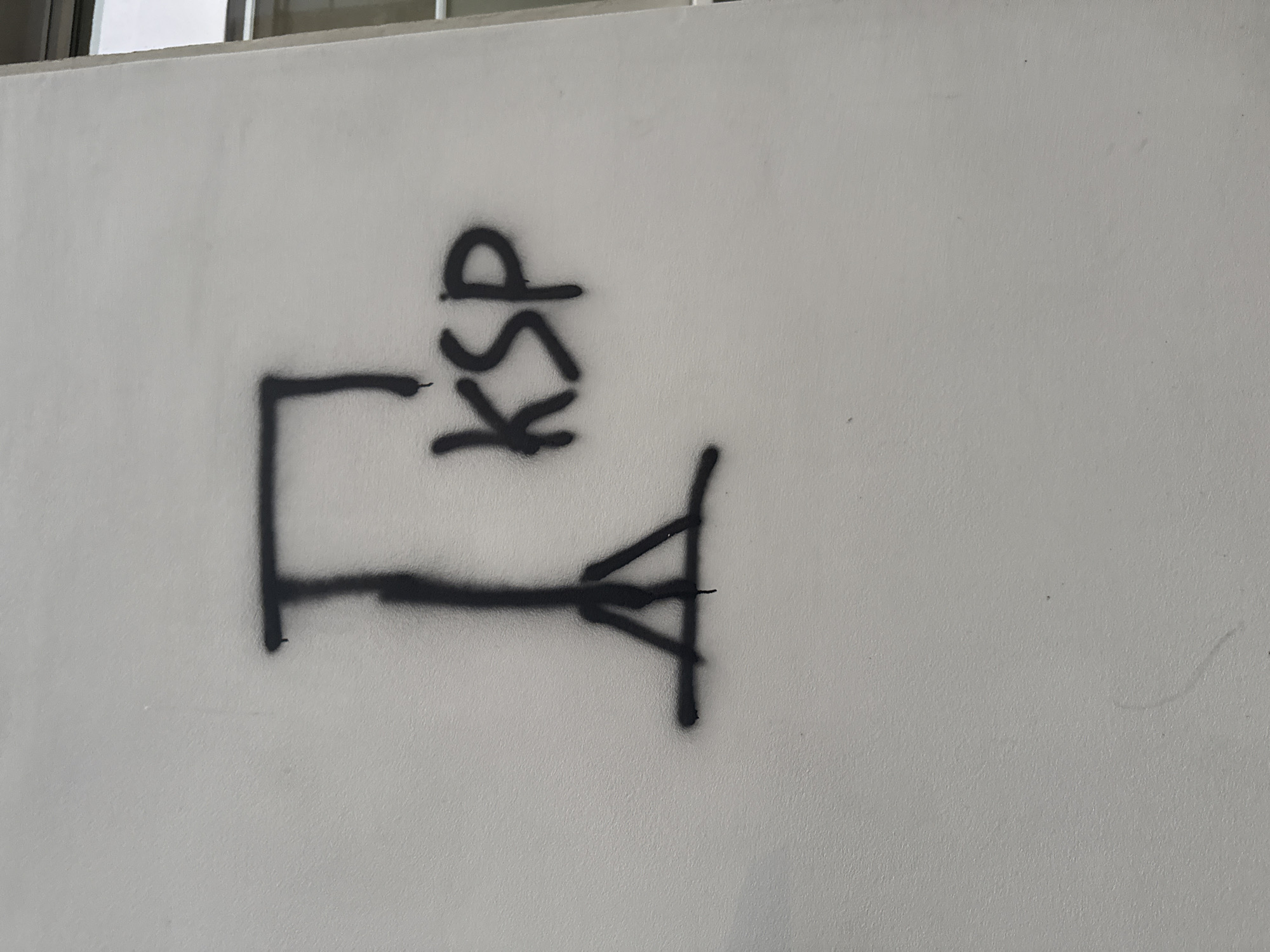 A black spray-painted symbol resembling a tripod with a horizontal bar extending from the top is depicted on a textured, light-colored wall. To the right of the symbol, the letters "KSP" are spray-painted in black. The background is a plain wall, with a sliver of a building with a window visible on the far right. The image is taken from a head-on perspective, at eye level.