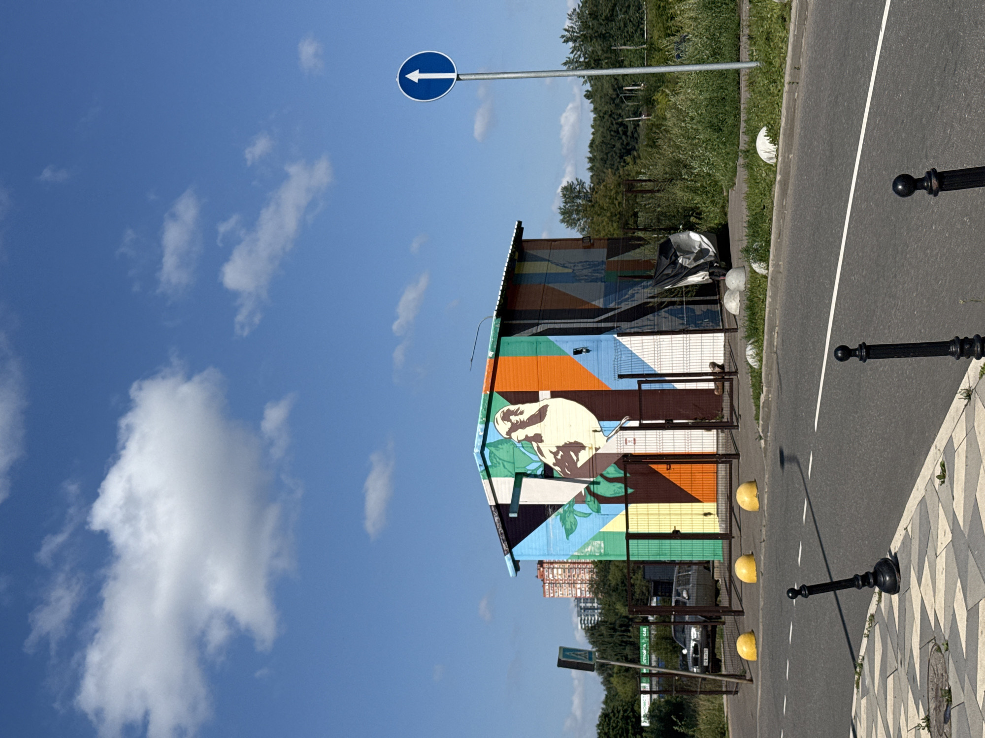 In Lyubertsy, Russia, on a sunny day with a bright blue sky and scattered white clouds, a small, rectangular building stands out with its vibrant, geometric mural. The mural features a large illustration of a bird, possibly a hawk or falcon, perched on branches with green leaves, set against a backdrop of colorful, abstract shapes in shades of orange, green, blue, and brown. The building appears to be a utility structure or storage unit, enclosed by a metal fence.
In front of the building, a paved road runs horizontally across the image. Yellow spherical bollards are placed along the edge of the road, separating it from a grassy area. Further down the road, a single blue traffic sign with a white upward-pointing arrow is mounted on a pole.
The foreground of the image shows a sidewalk with a distinctive patterned paving. Two black, decorative bollards are positioned on the sidewalk. In the background, beyond the fence and grassy area, a dense line of trees forms a green backdrop, with several multi-story residential buildings visible further in the distance. A silver car is parked behind the fence to the left of the building. The scene suggests an urban or suburban environment on a clear day.