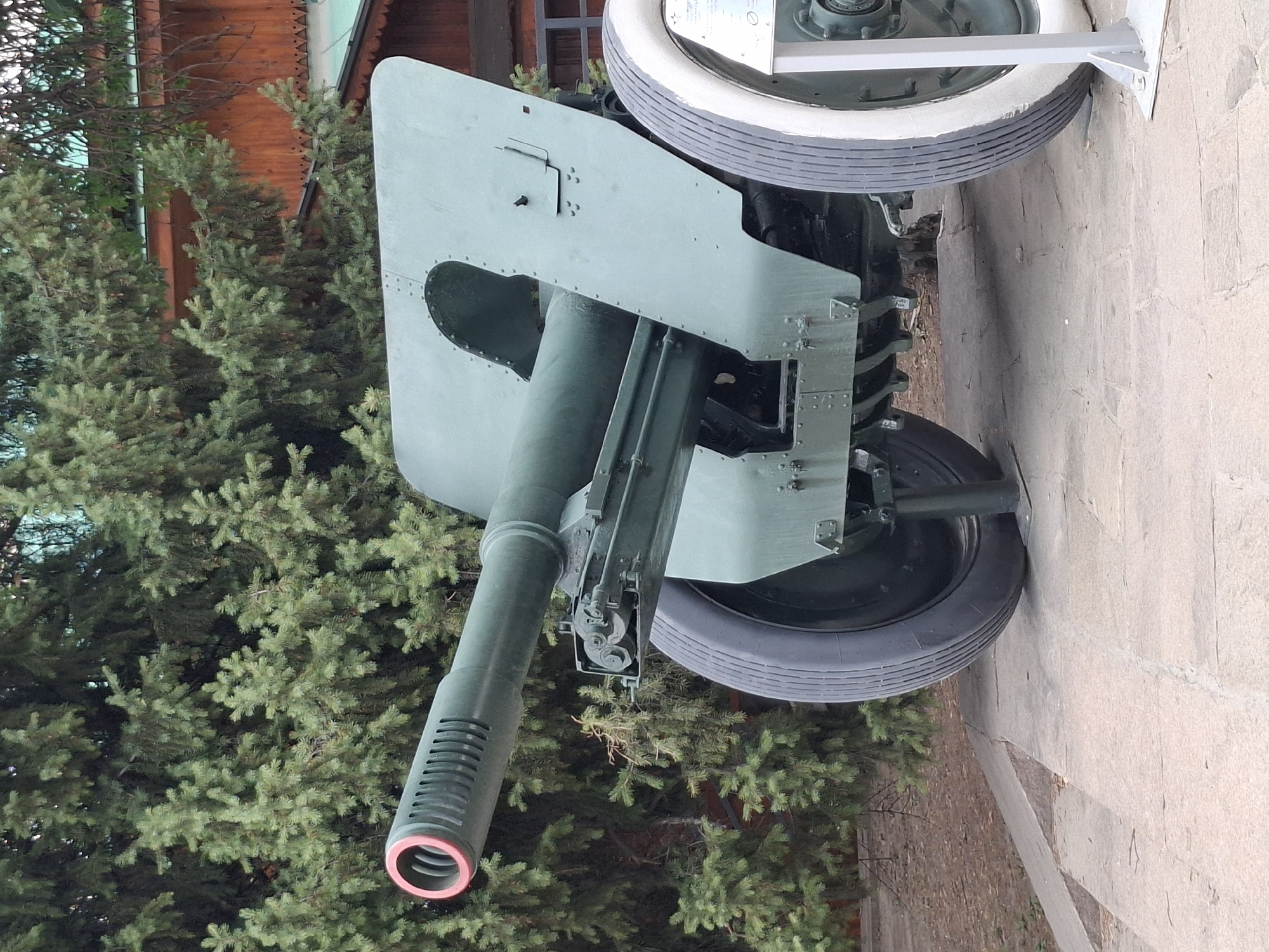 A large, olive-green artillery piece is positioned outdoors on a paved surface. The cannon is angled slightly to the right, with its barrel extending forward and featuring a ribbed muzzle brake with a red ring. The piece is mounted on a carriage with two large wheels, the one on the right being notably larger and having a white rim and a dark tire. The background consists of dense green evergreen trees and a wooden building with a dark roof. The scene appears to be in a park or museum setting.