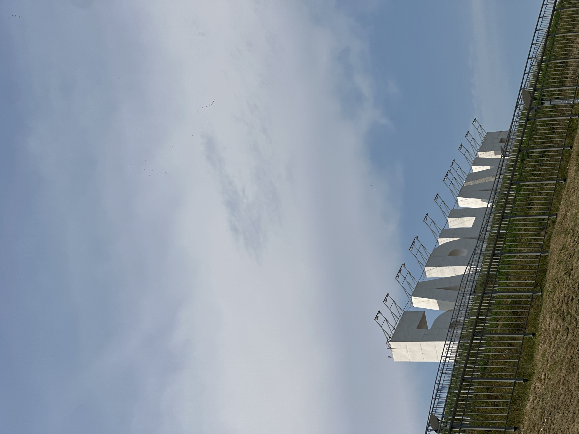 The image shows a large, white sign with the letters "БАРНАУЛ" (Barnaul) prominently displayed against a blue sky with scattered clouds. The sign is situated on a grassy embankment behind a metal fence. The letters are three-dimensional and appear to be made of concrete or a similar material. They are illuminated by lights mounted on metal structures above them. The grassy slope is dry and appears to be covered with sparse vegetation. There are no people visible in the image. The scene is a wide shot, capturing the sign and its immediate surroundings. The image was taken outdoors, likely during the daytime.
