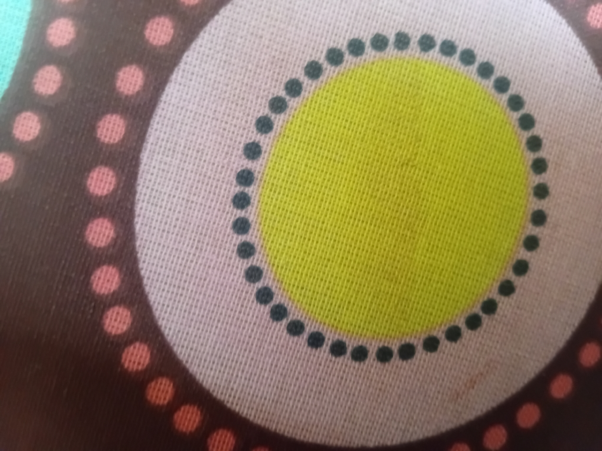 The image shows a close-up of a patterned fabric. The pattern features concentric circles. The innermost circle is bright yellow, surrounded by a pale pink or cream-colored ring. This ring is bordered by a dark brown band with a pattern of reddish-pink dots. Inside the pale ring and just outside the yellow circle, there is a band of dark grey or black dots. The fabric appears to be textured, likely a woven material like cotton or linen. There are no people, actions, or interactions visible. The image is an abstract detail of a textile design and does not provide any information about its location in Verkhniachka, Ukraine, or any other geographical context.