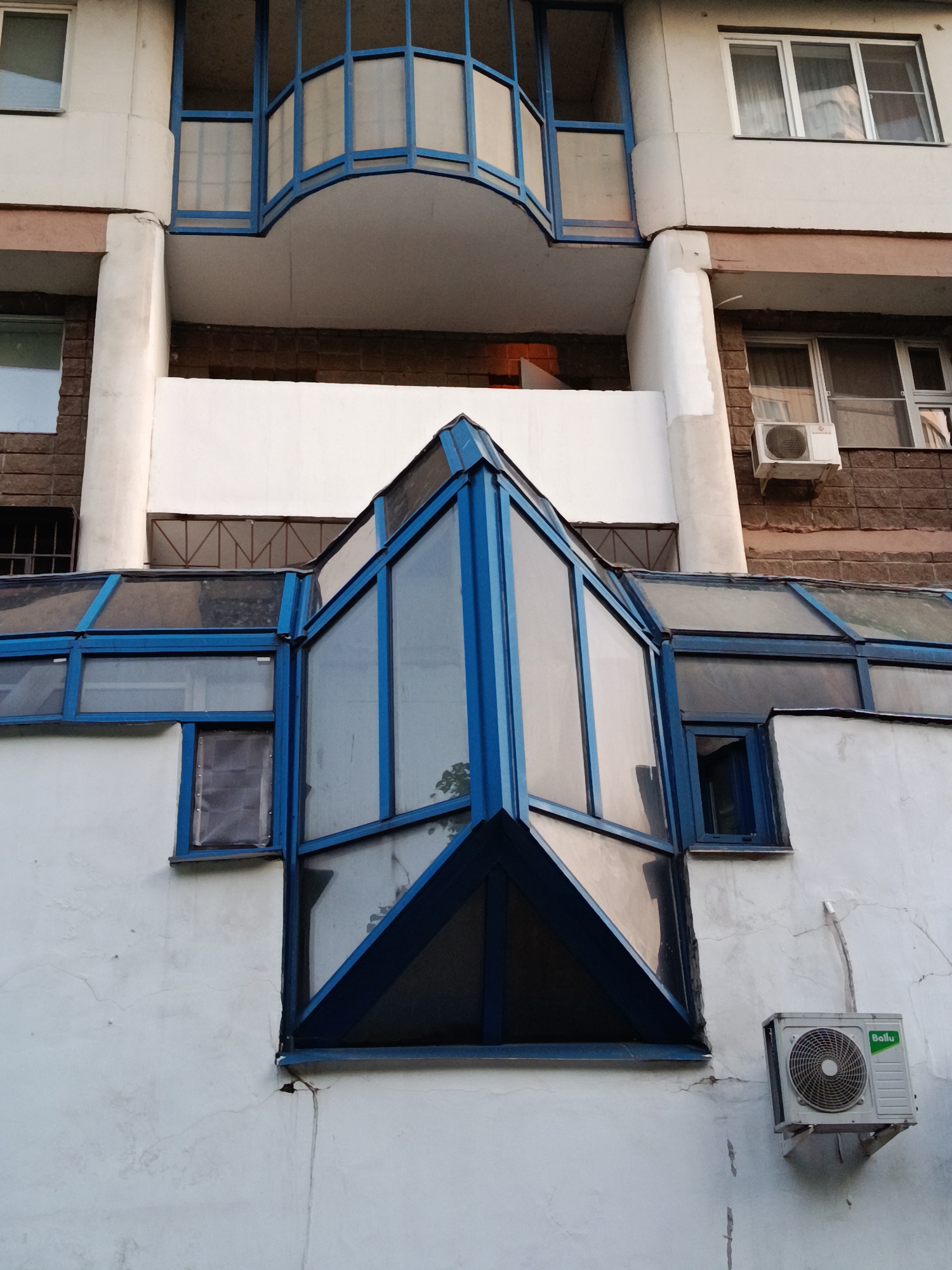 This is an exterior shot of an apartment building with a mix of architectural styles located in Moscow, Russia. The structure features a prominent blue-framed glass bay window on the lower portion, adjoining what appears to be a greenhouse-like extension with a similar blue frame. The building's facade is a mix of white and brown brick, with columns and a curved, enclosed balcony on an upper level. An air conditioning unit is mounted on the lower section of the building to the right of the bay window. The overall impression is of a building constructed or modified over time with varying styles. The building appears to be aged, and shows signs of wear and tear.
