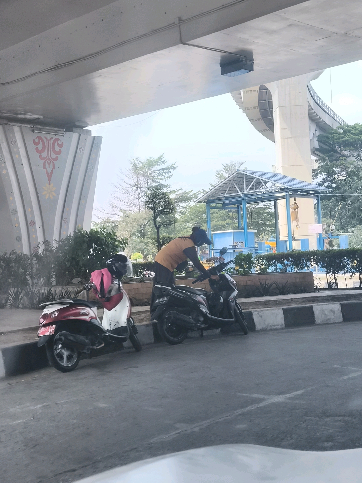 The image captures a scene under an elevated road structure, likely part of the Palembang Light Rail Transit (LRT) infrastructure, given the specific architectural style of the supporting pillars and the overall urban environment. In the foreground, two scooters are parked on the side of a road. The scooter on the left is red and white, with a pink garment draped over its handlebars and a helmet. The scooter on the right is black. A person wearing an orange shirt and a dark cap is leaning over the black scooter, appearing to interact with it. The background features lush green vegetation, a blue canopy structure with a crane, and the imposing concrete pillars of the elevated road. The overall ambiance suggests a daytime scene in an urban area of Palembang, Indonesia.