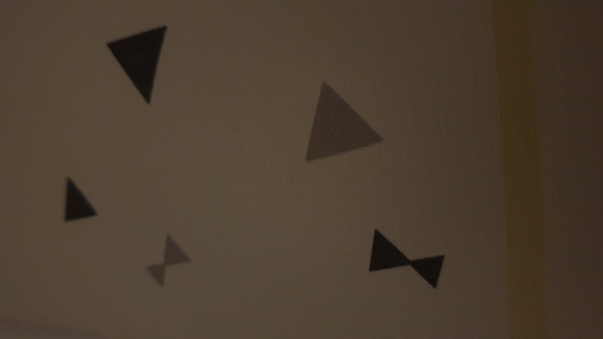The image shows a patterned surface, possibly a wall or fabric, with several dark geometric shapes resembling triangles and a bow-tie shape. These shapes are scattered across the surface at various orientations and sizes. The lighting in the image is dim, casting shadows and making the details of the pattern less distinct. The surface itself appears to have a subtle texture, possibly a woven material or a textured paint. There is a horizontal band of lighter color, likely a transition to another surface or a painted stripe, at the bottom of the image. The scene does not contain any people, actions, or interactions. There is no information within the image that suggests a specific location in Vrchotovy Janovice, Czechia, or any other geographical context.