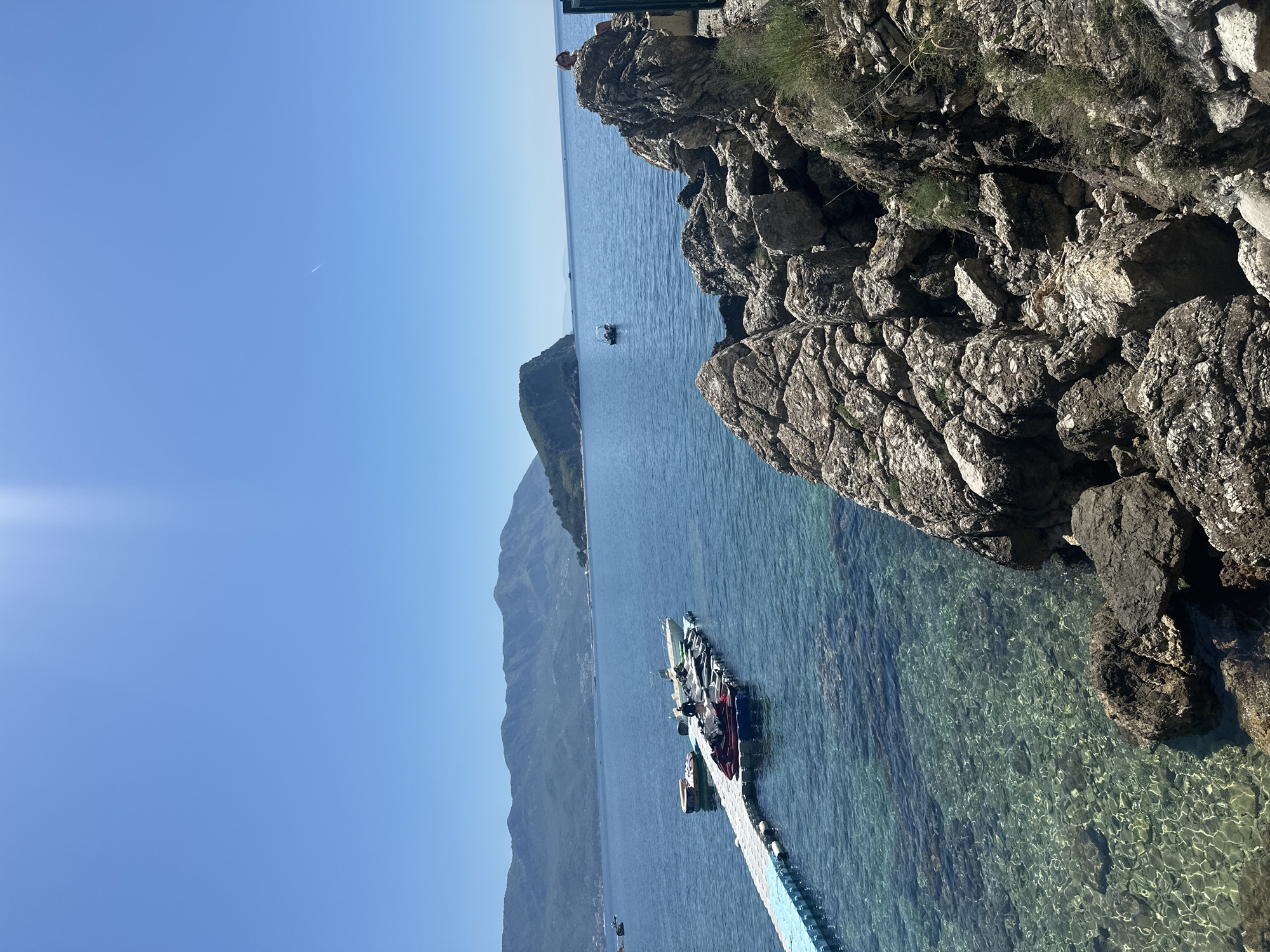The image presents a coastal scene, likely taken in Budva, Montenegro. The view is dominated by the Adriatic Sea, where crystal-clear water reveals the seafloor. A floating dock extends into the sea, holding multiple jet skis and boats. In the background, a mountain range stretches along the coastline. Rocky cliffs, covered with sparse vegetation, are visible on the right side of the frame. The sky is clear blue with a jet plane visible on the upper left.